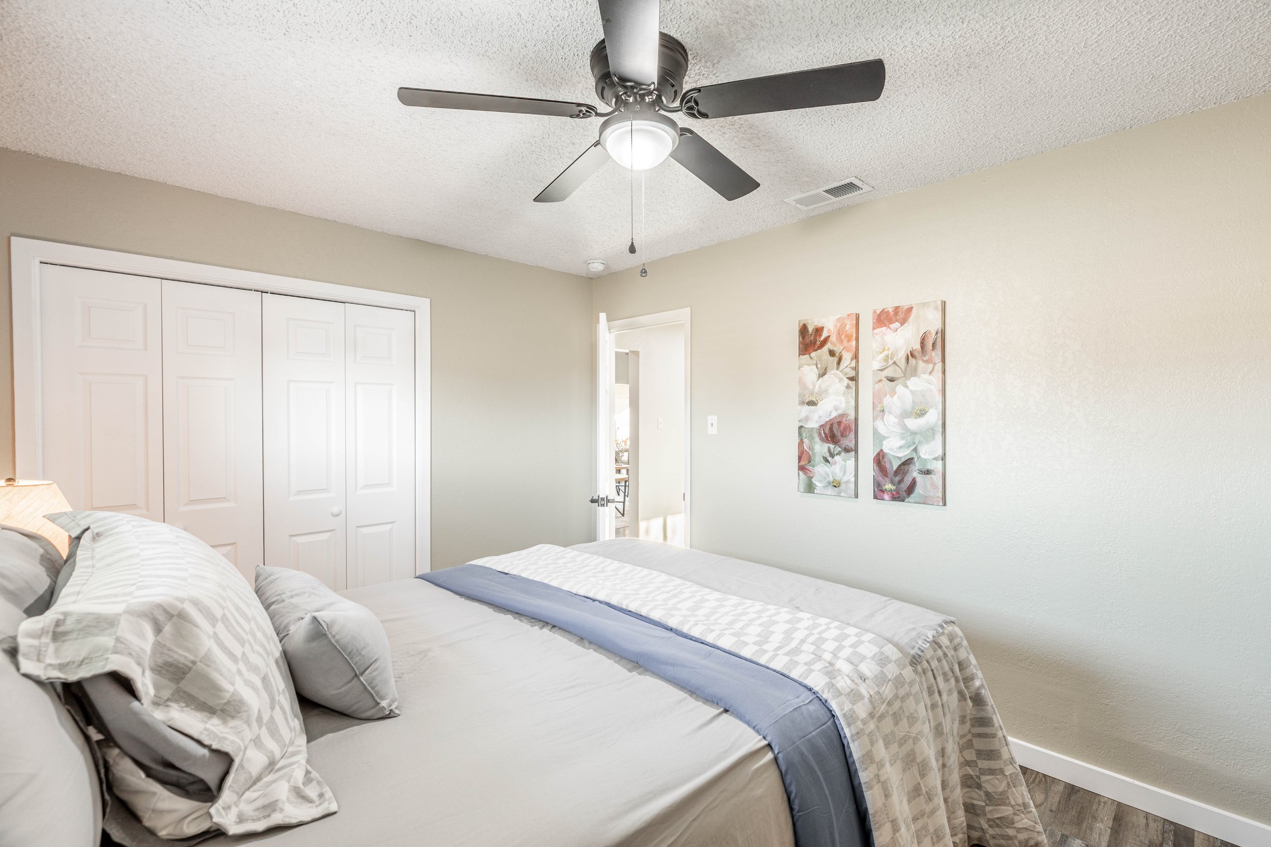 A bedroom with a ceiling fan, white walls, a closet with white doors, and floral artwork on the wall near the bed.