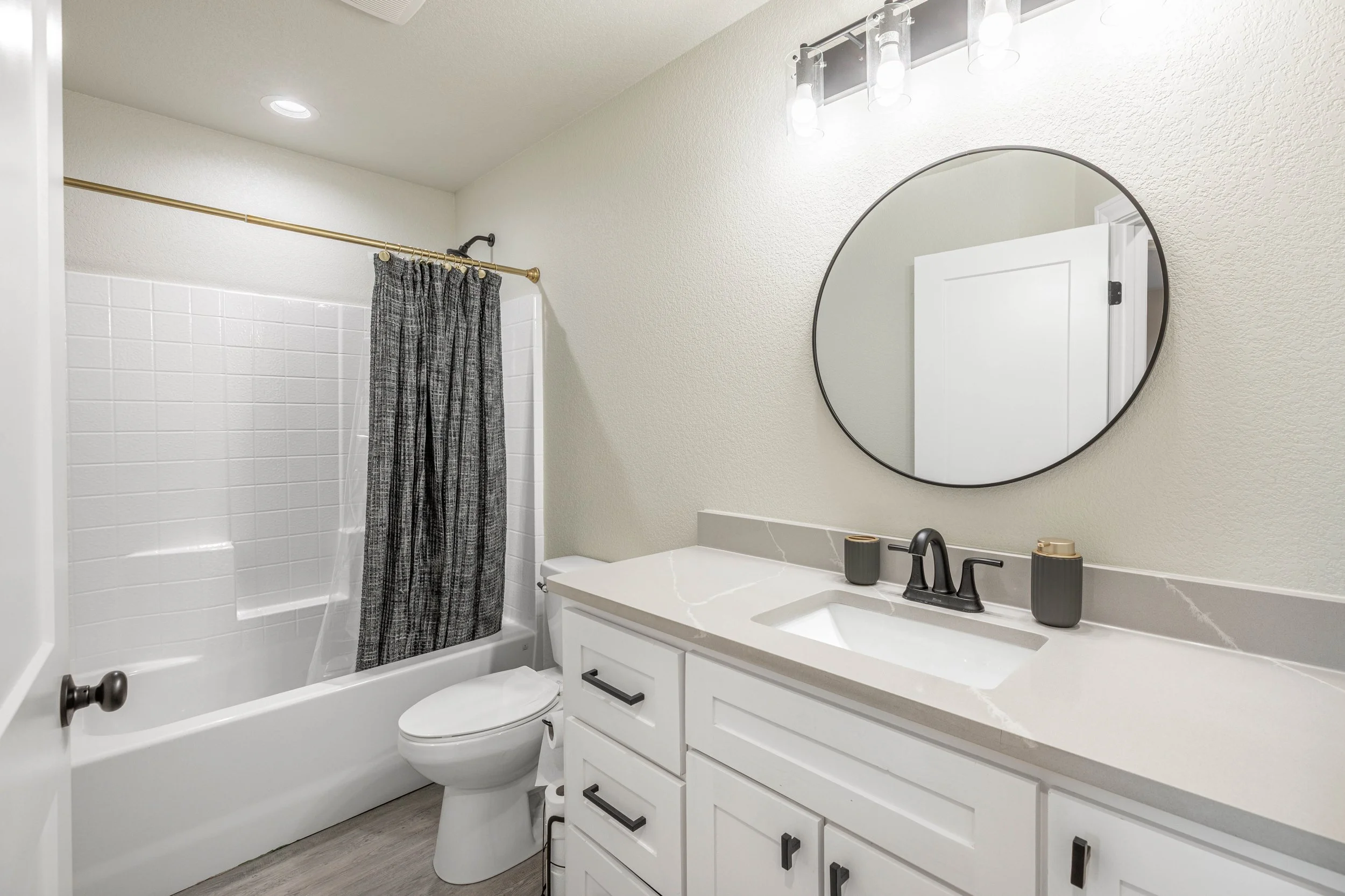 A modern bathroom with a white vanity, black fixtures, a round mirror, and a white bathtub with a gray and black curtain.