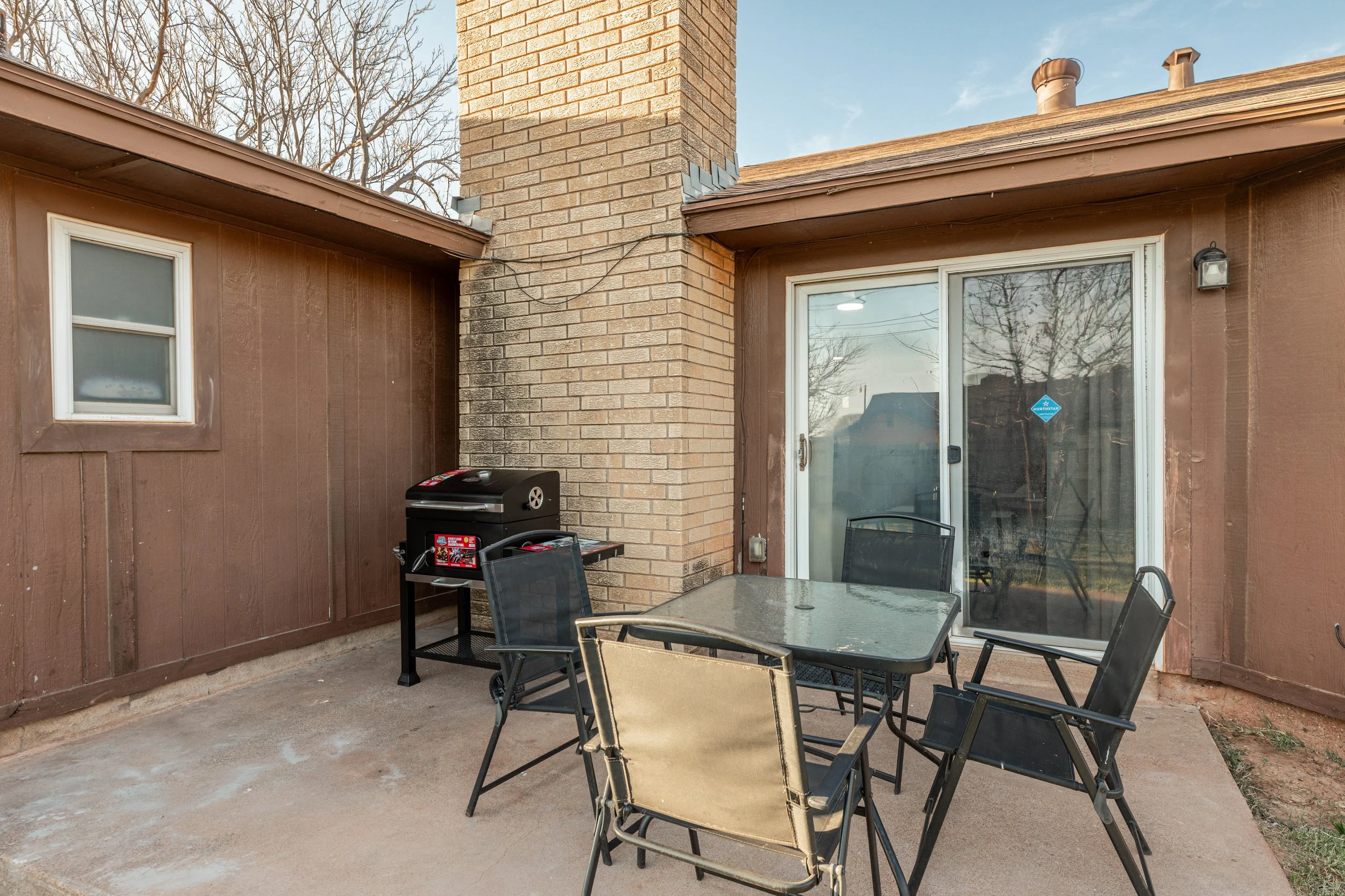 Back patio with brown wooden siding, a brick chimney, a sliding glass door, outdoor table and chairs, and a black grill