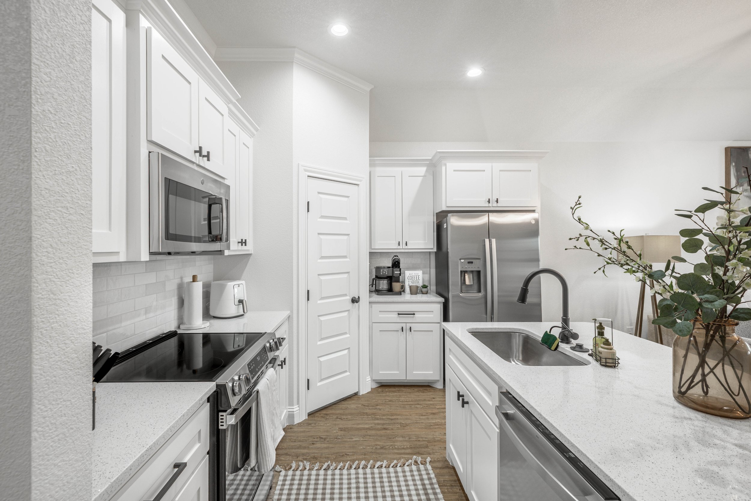 Modern white kitchen with stainless steel appliances, including a refrigerator and microwave, white cabinets, and a white subway tile backsplash. Countertop with various items and a large vase with greenery.