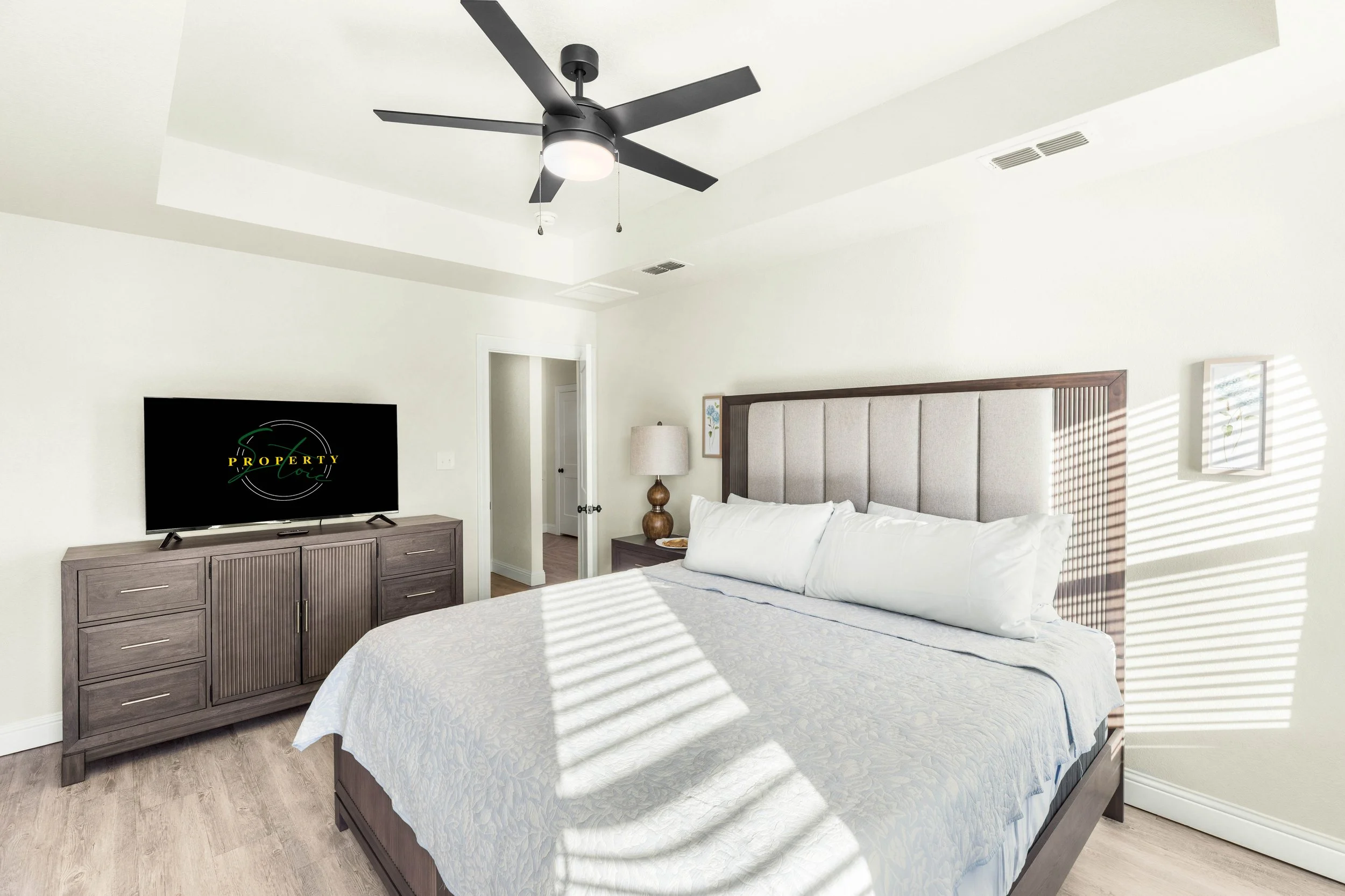 A bedroom with a bed having a beige upholstered headboard, white bedding, and a gray wooden dresser with a television on top. There is a lamp next to the bed and framed artwork on the wall. Sunlight creates striped shadows through window blinds.