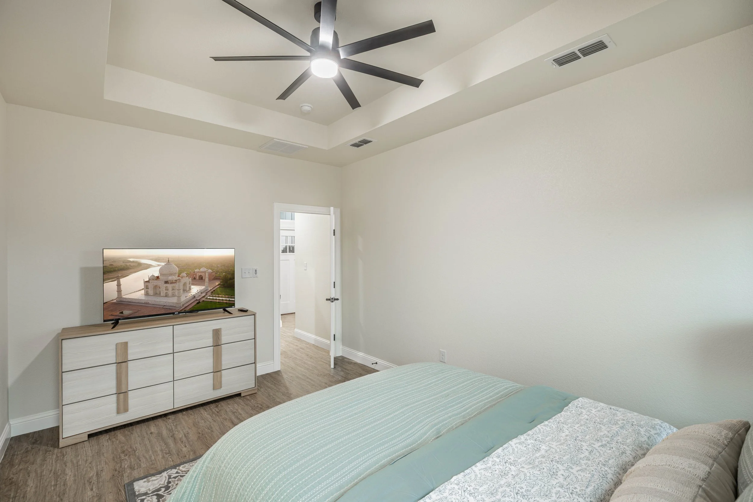 A bedroom with a flat-screen TV on a white dresser, a bed with light green and beige linens, a ceiling fan, and white walls.