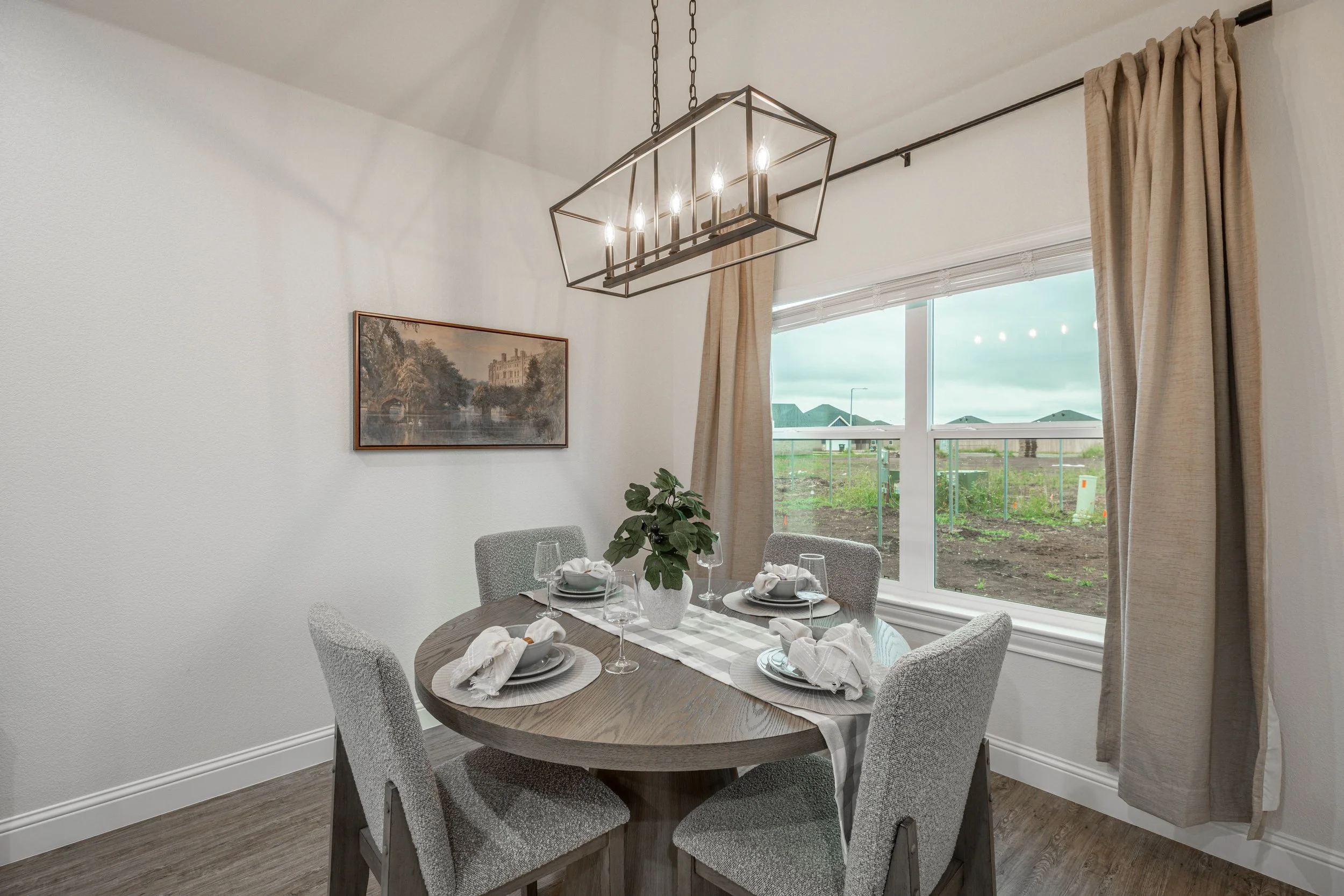 A dining room with a round wooden table set for four, decorated with a gray and white table runner, white plates, napkins, wine glasses, and a white potted plant centerpiece. Four upholstered chairs surround the table. A large window with beige curta