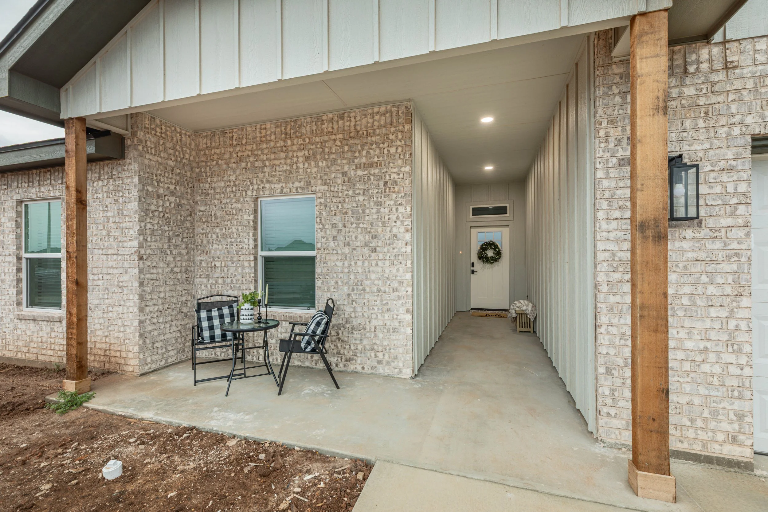 Front porch area of a house with a brick exterior, small round table with four chairs, and decorative wreath at the doorway. Wooden posts support the roof, and there are two windows and a lantern-style light fixture.