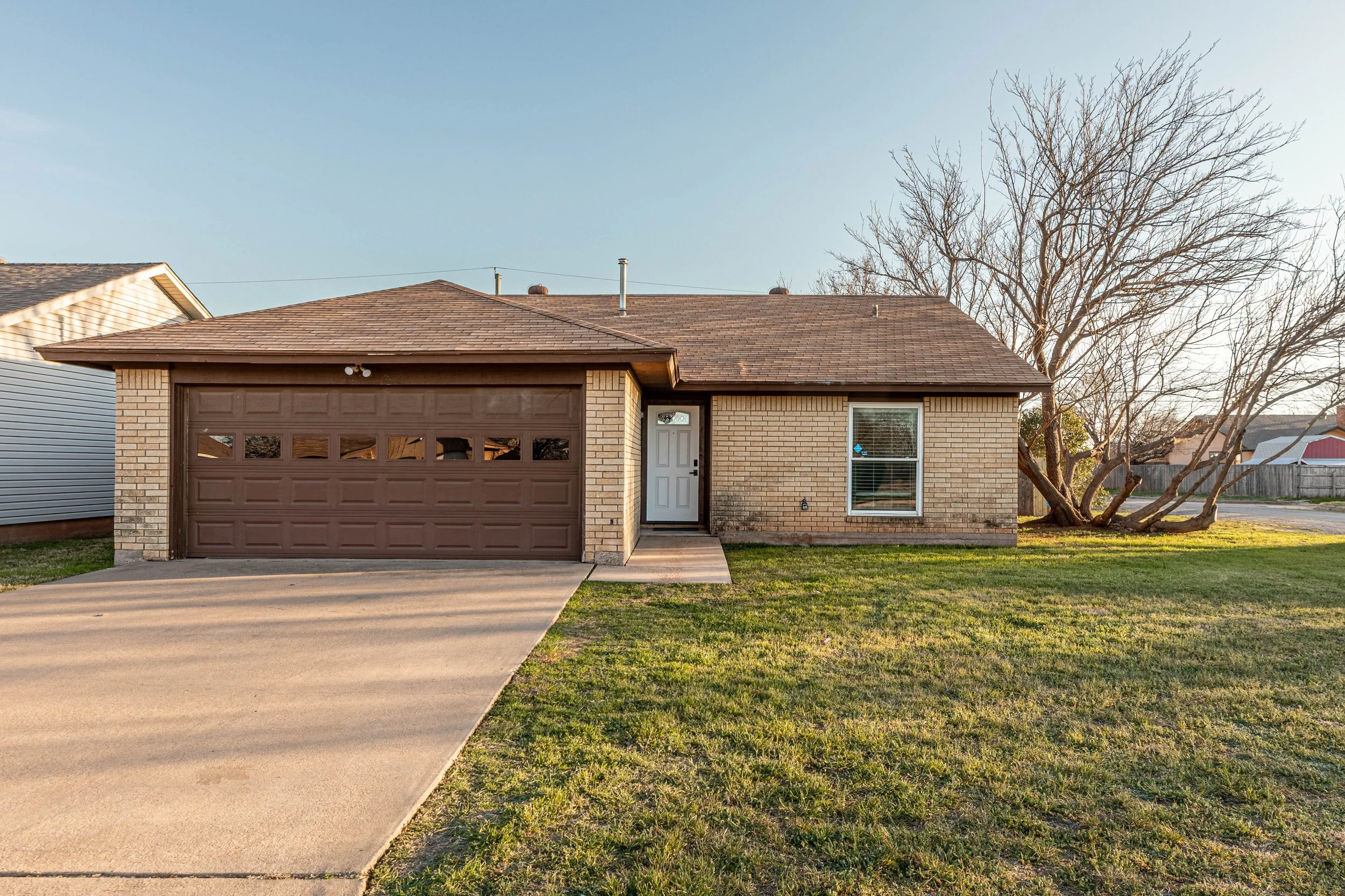 Front view of a brick house with a brown garage door, white front door, single window, and a large, leafless tree on the right side. The house is in a suburban neighborhood with neighboring houses and a clear blue sky.