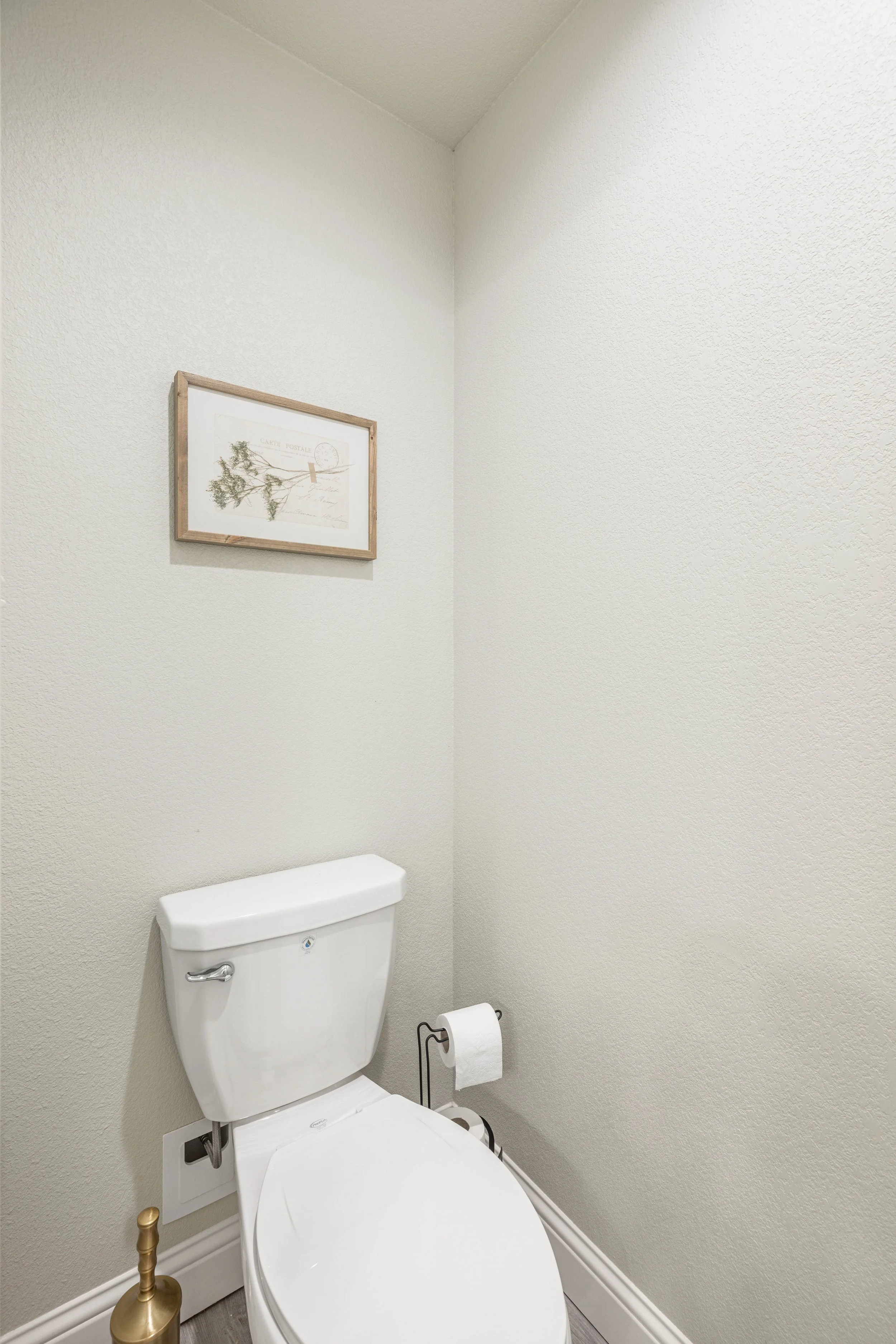 A small bathroom with beige textured walls, a white toilet, a framed piece of artwork above it, a toilet paper roll on a black holder, and a gold-colored toilet brush.