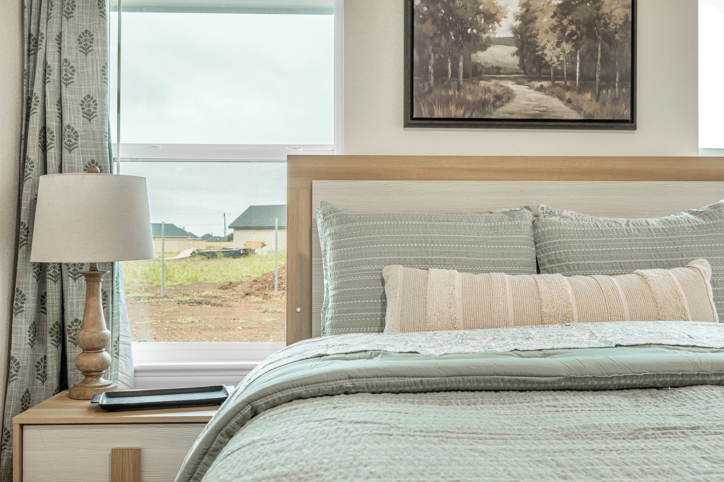 A bedroom with a bed covered in light green bedding, two pillows, a nightstand with a lamp, a window with curtains, and a painting of a wooded landscape on the wall.
