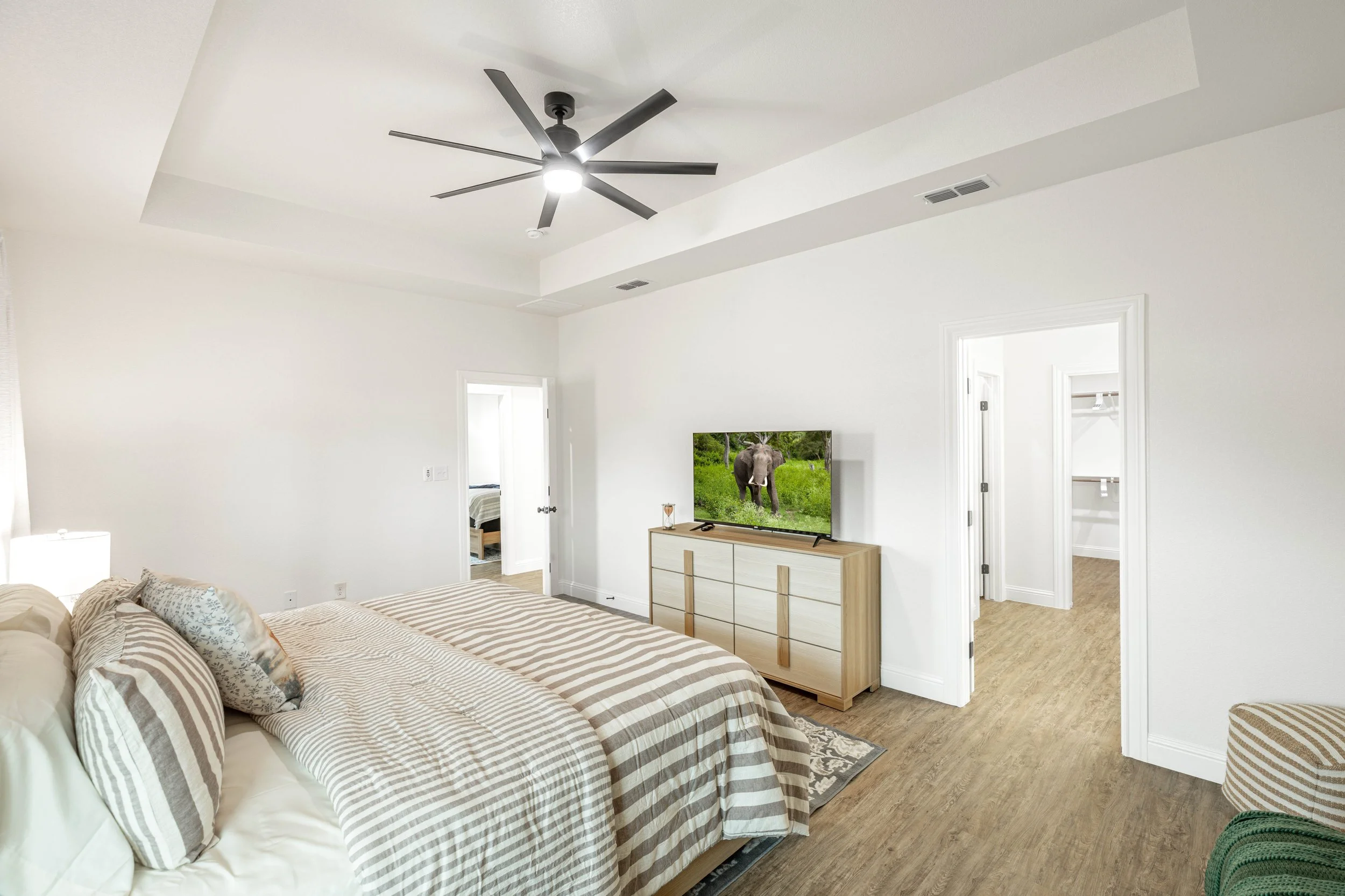 A bedroom with a bed covered in beige and white striped bedding and pillows. A wooden dresser with a TV displaying an elephant, near White walls and a ceiling fan, with open doors leading to other rooms.