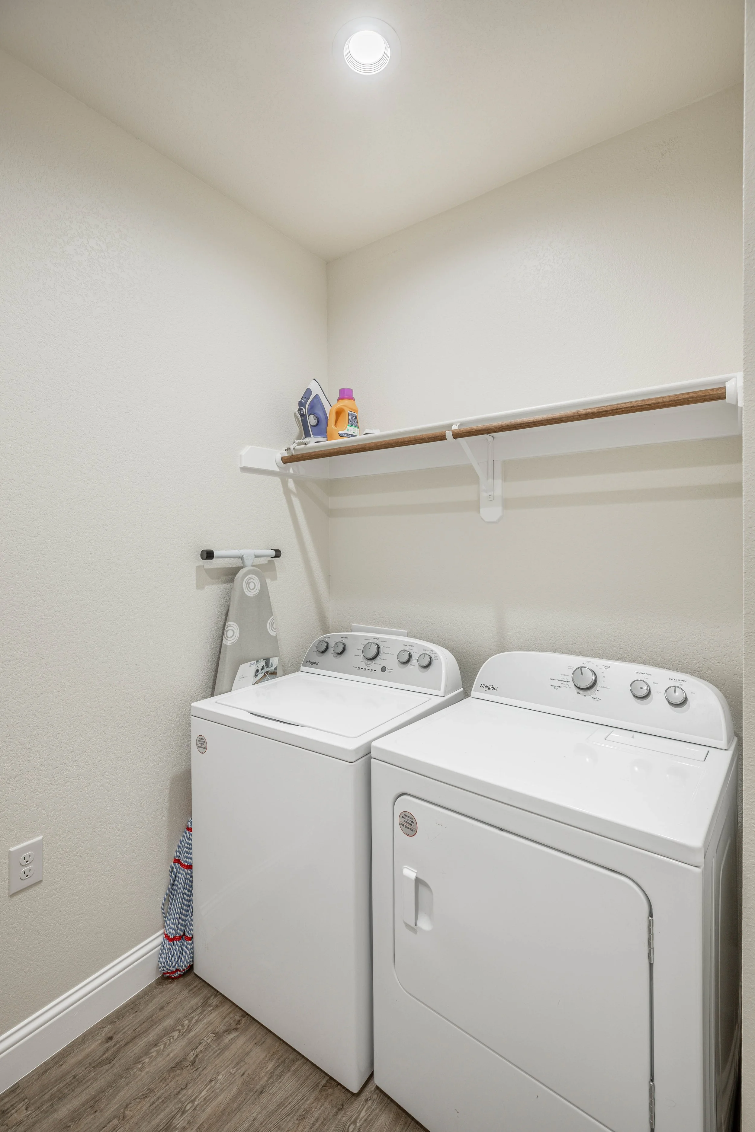 A laundry room with a washing machine and dryer, a wooden shelf holding laundry supplies, an ironing board, and a striped cloth hanging, with a ceiling light overhead and wood-look flooring.