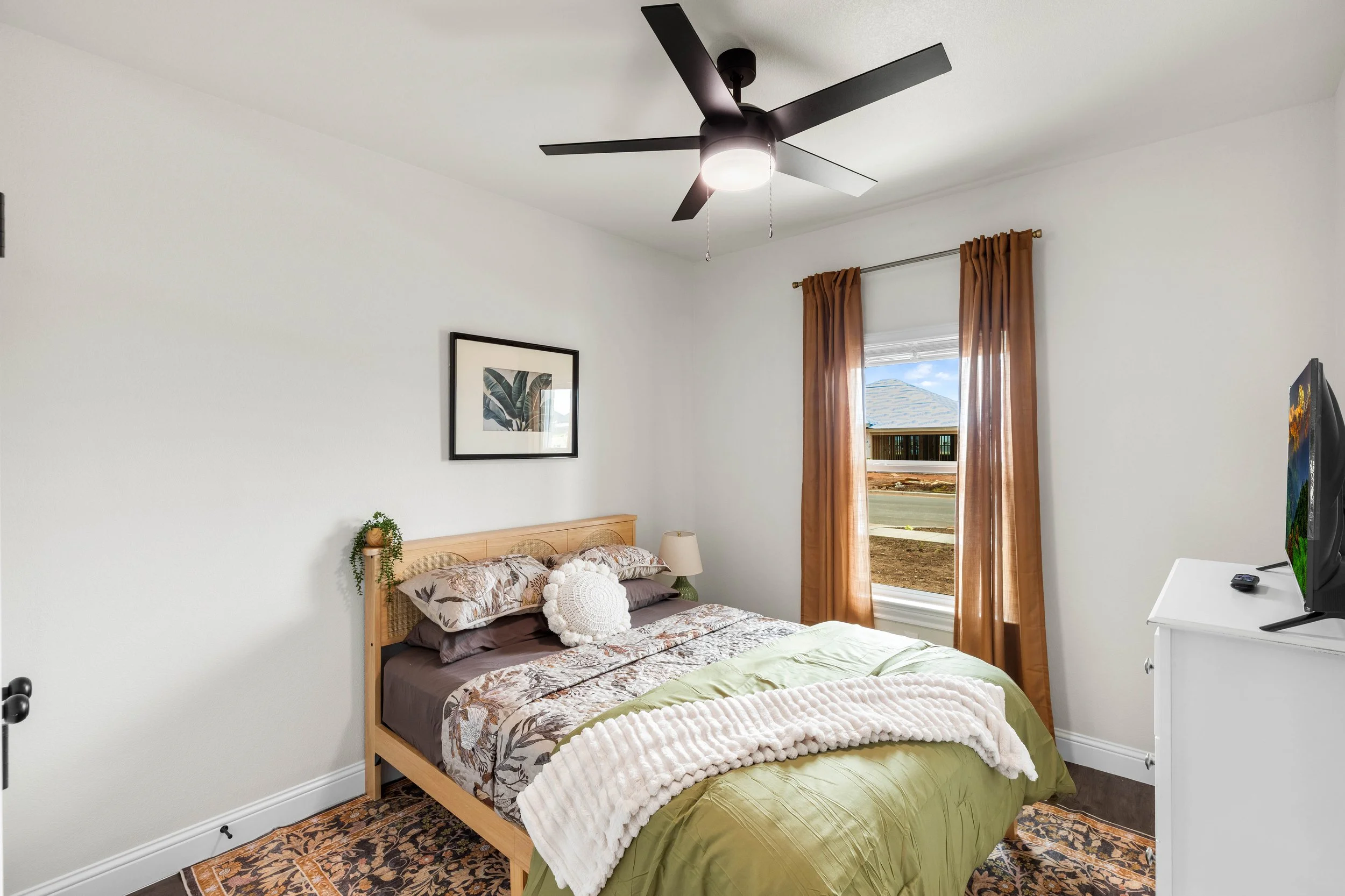 A bedroom with a bed, artwork on the wall, a ceiling fan, a window with curtains, and a dresser with a TV on it.