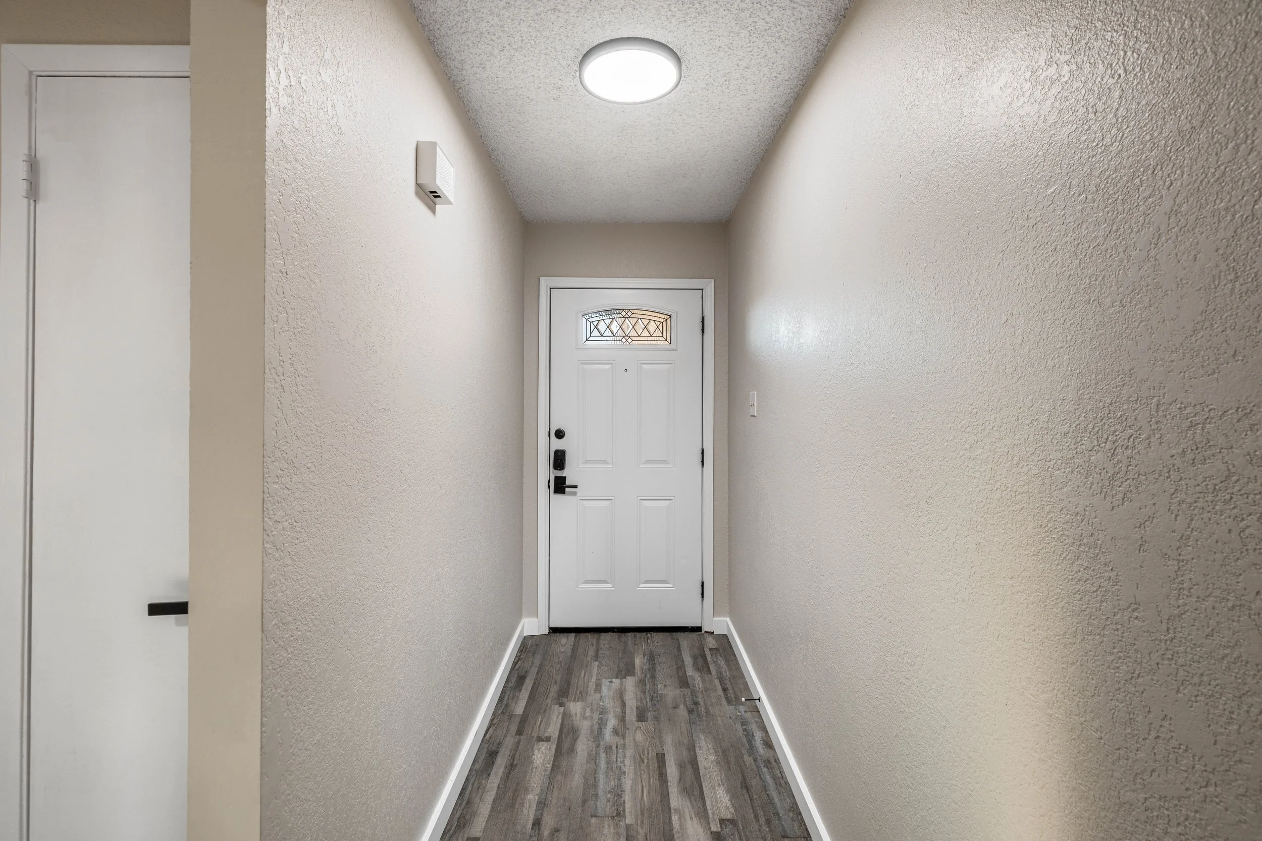 Interior hallway with beige textured walls, a white front door with decorative glass, a ceiling light, and wood planked flooring.