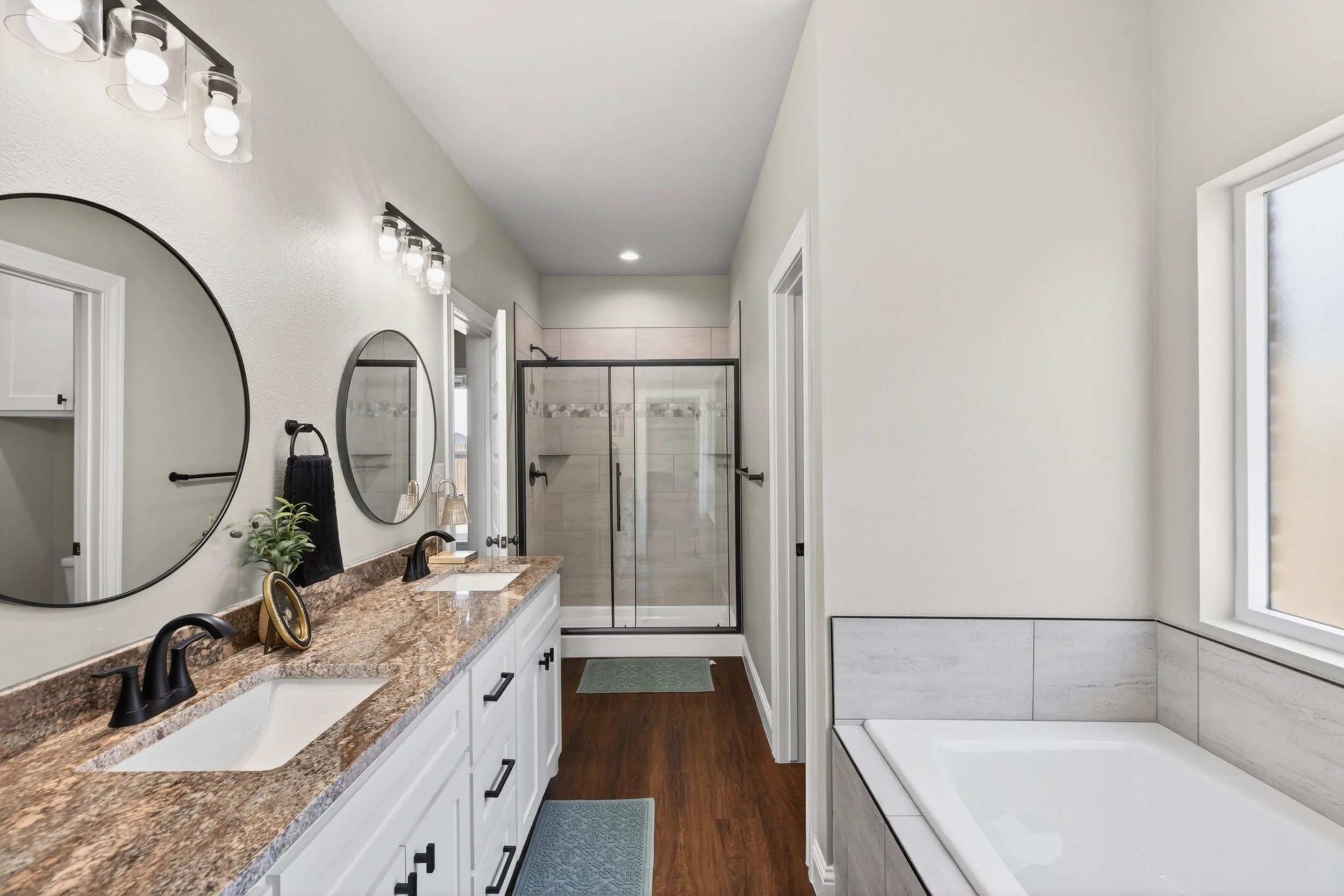A modern bathroom with a double vanity, black fixtures, oval mirrors, and a tiled shower with a glass door. There is a bathtub in the corner and a window allowing natural light.