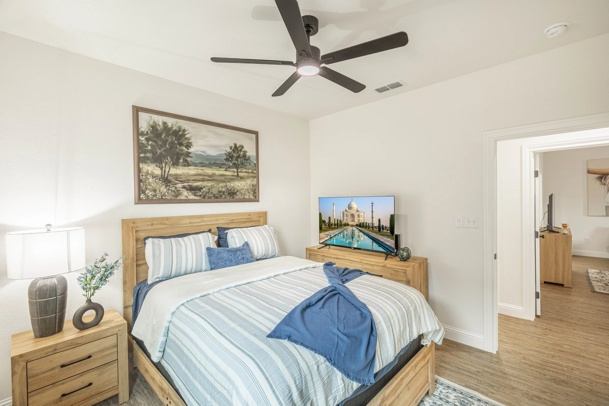 A bedroom with a wooden bed frame, striped bedding, and blue pillows. A nightstand with a modern vase and flowers. A landscape painting on the wall above the bed. A flat-screen TV displaying the Taj Mahal in the background. A ceiling fan with five da
