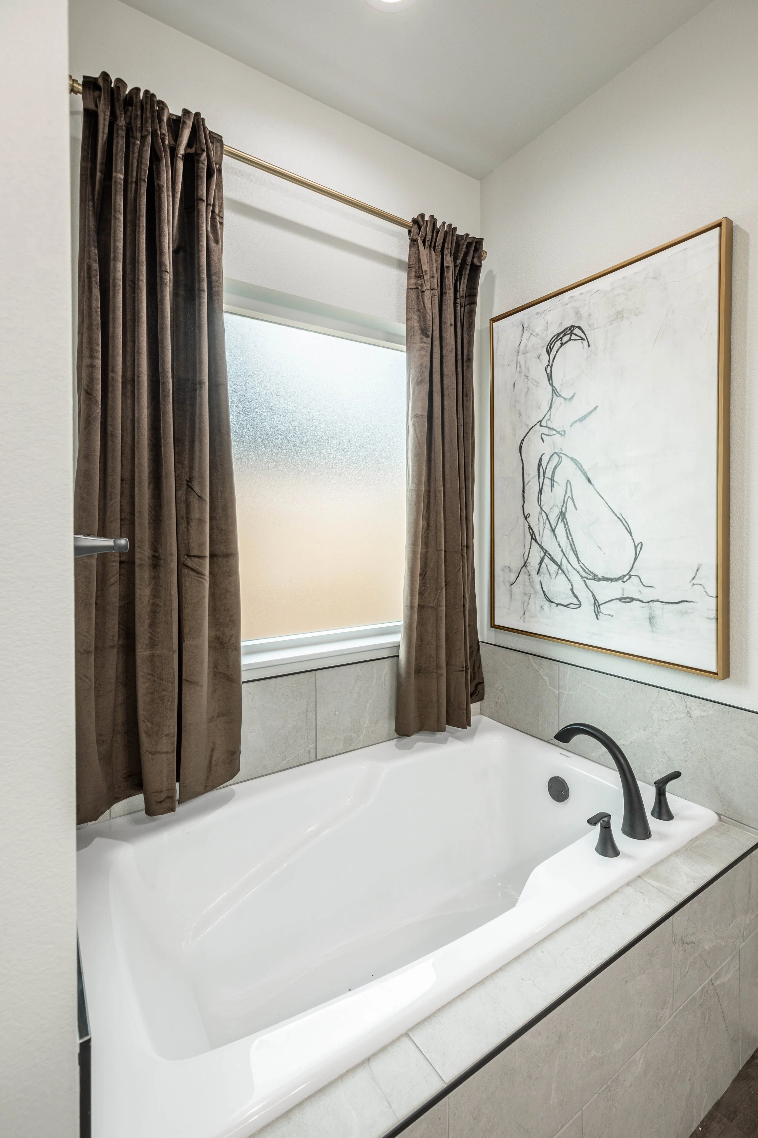 A bathroom with a white bathtub, black faucet, and dark brown curtains. There is a large framed art piece with a minimalist line drawing of a person sitting, hanging on the wall above the tub. The room has white walls and gray marble floor tiles.
