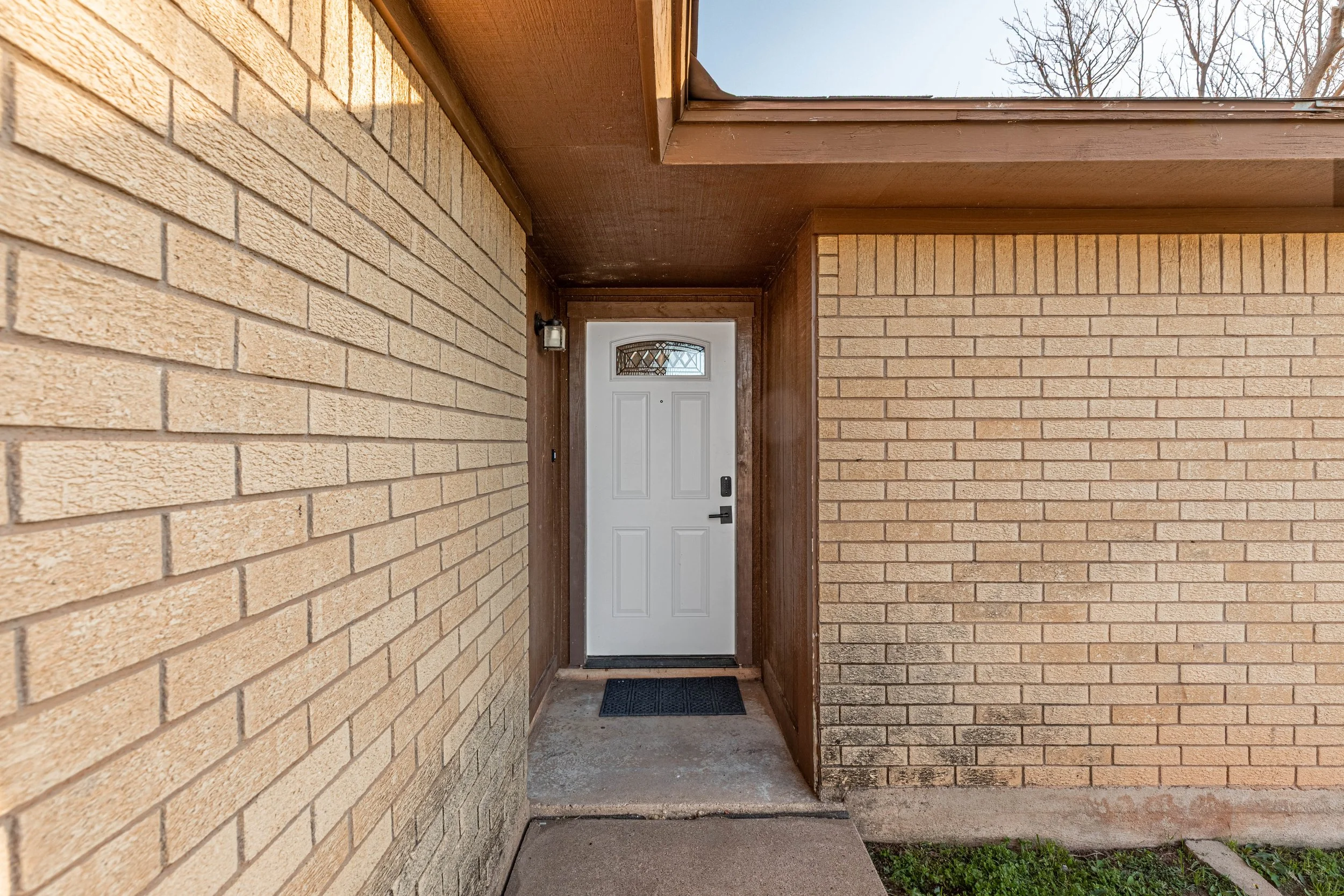 This image shows a small covered entryway with beige brick walls on both sides and a white front door with a decorative window at the top. There is a small black outdoor light fixture next to the door, and a black doormat on the concrete floor. The s