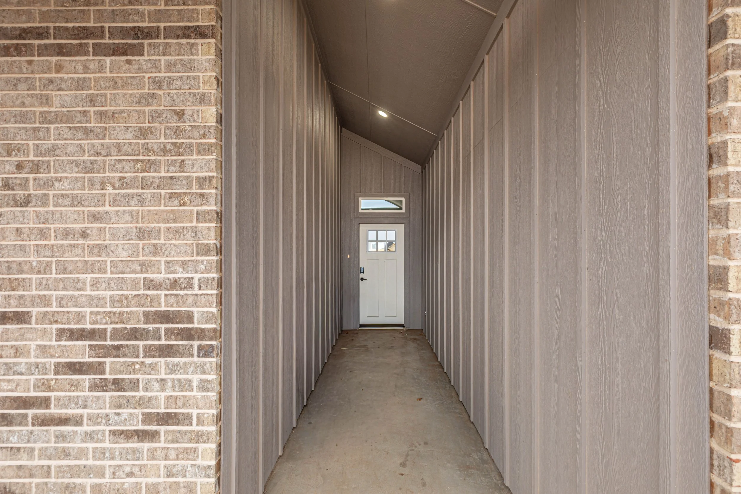 A narrow hallway with brick and wood-paneled walls, a concrete floor, and a white door with small windows at the far end.