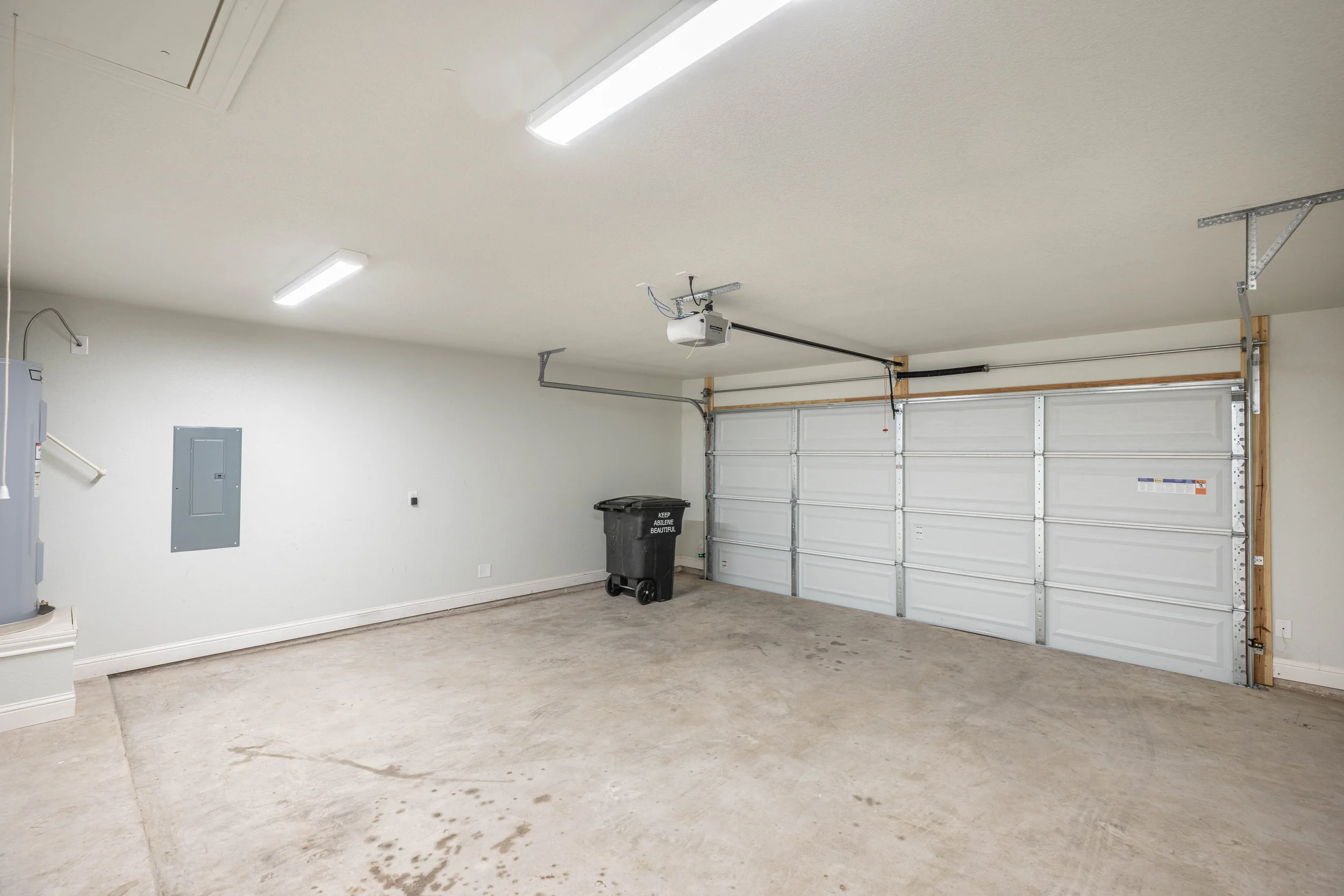 A clean, empty garage with a white ceiling, fluorescent lights, a garage door with metal panels, and a trash bin in the corner. The floor is concrete with some stains.