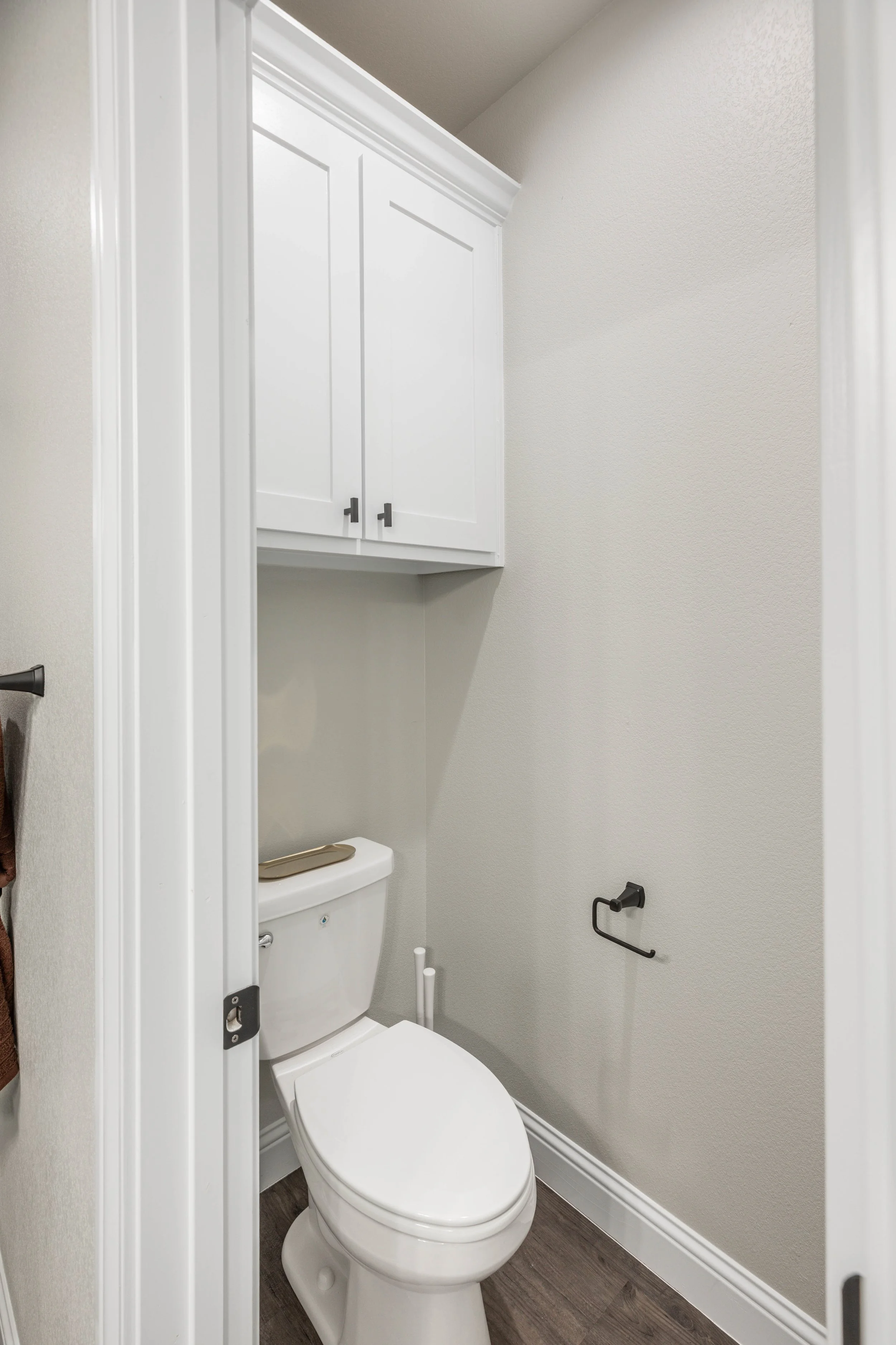 Small bathroom with a toilet, white cabinet above, gray walls, toilet paper holder on the wall, and dark wood flooring.