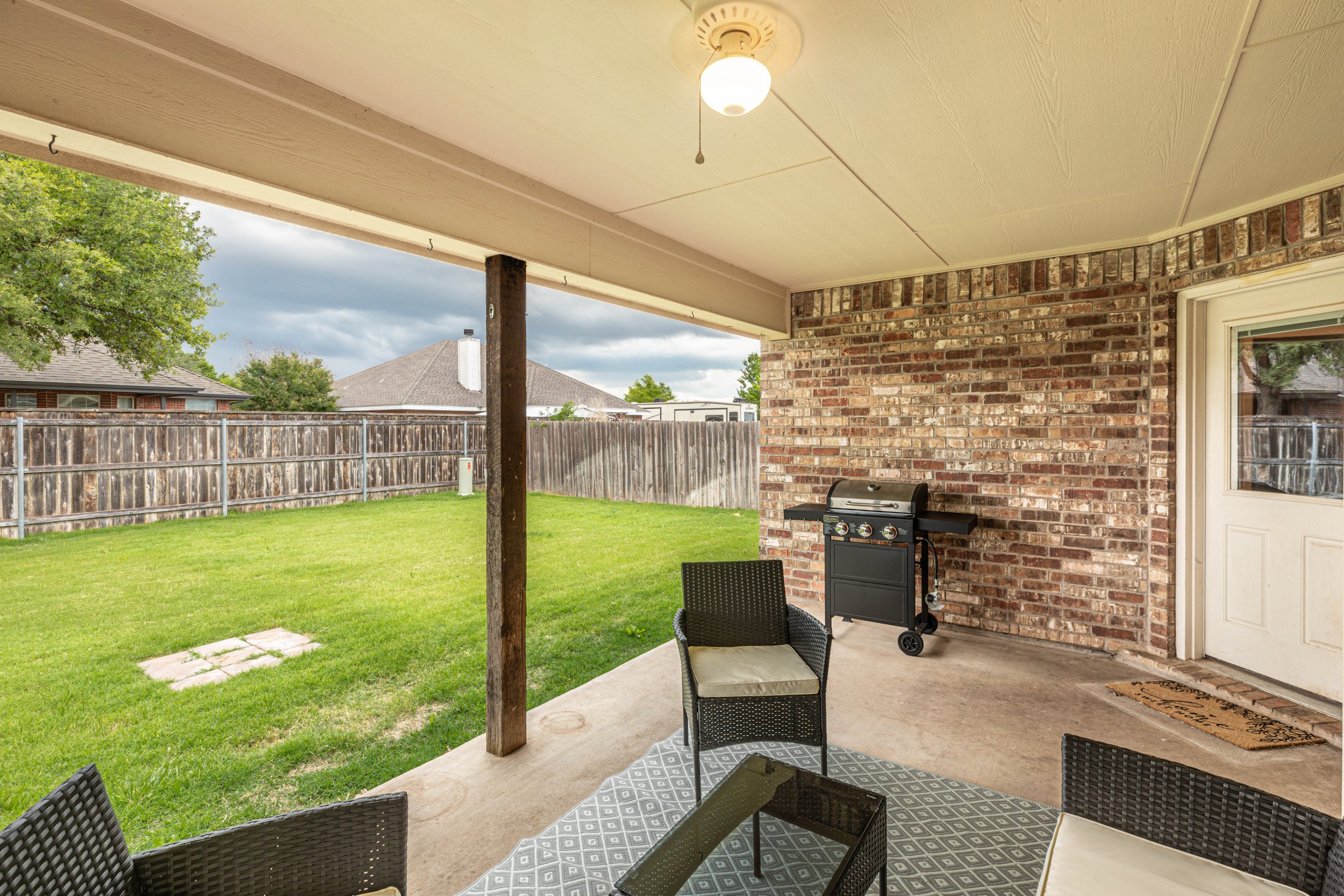 A covered patio area with outdoor furniture, a barbecue grill, and a view of a backyard with grass and a wooden fence.