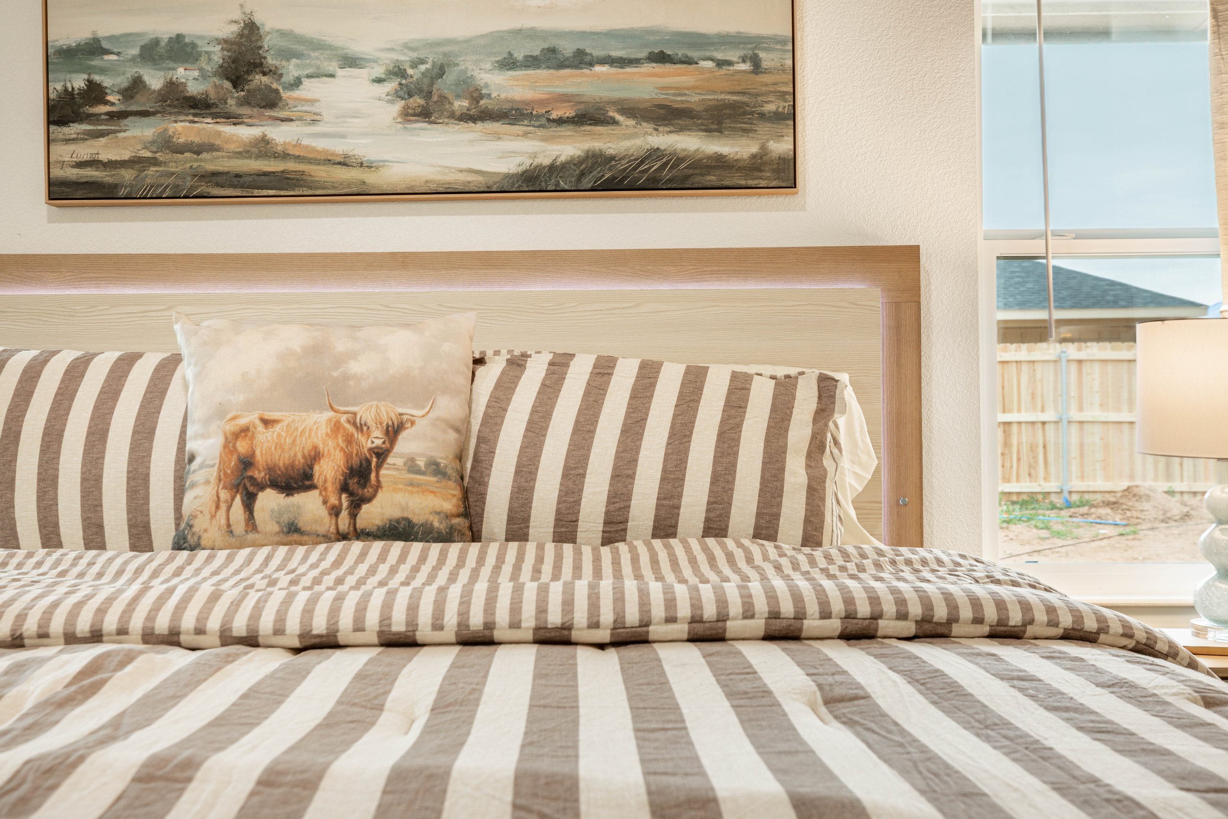 Bedroom with bed, striped bedding, pillow with cow picture, abstract landscape painting above bed, window with blinds showing outdoor fenced yard.