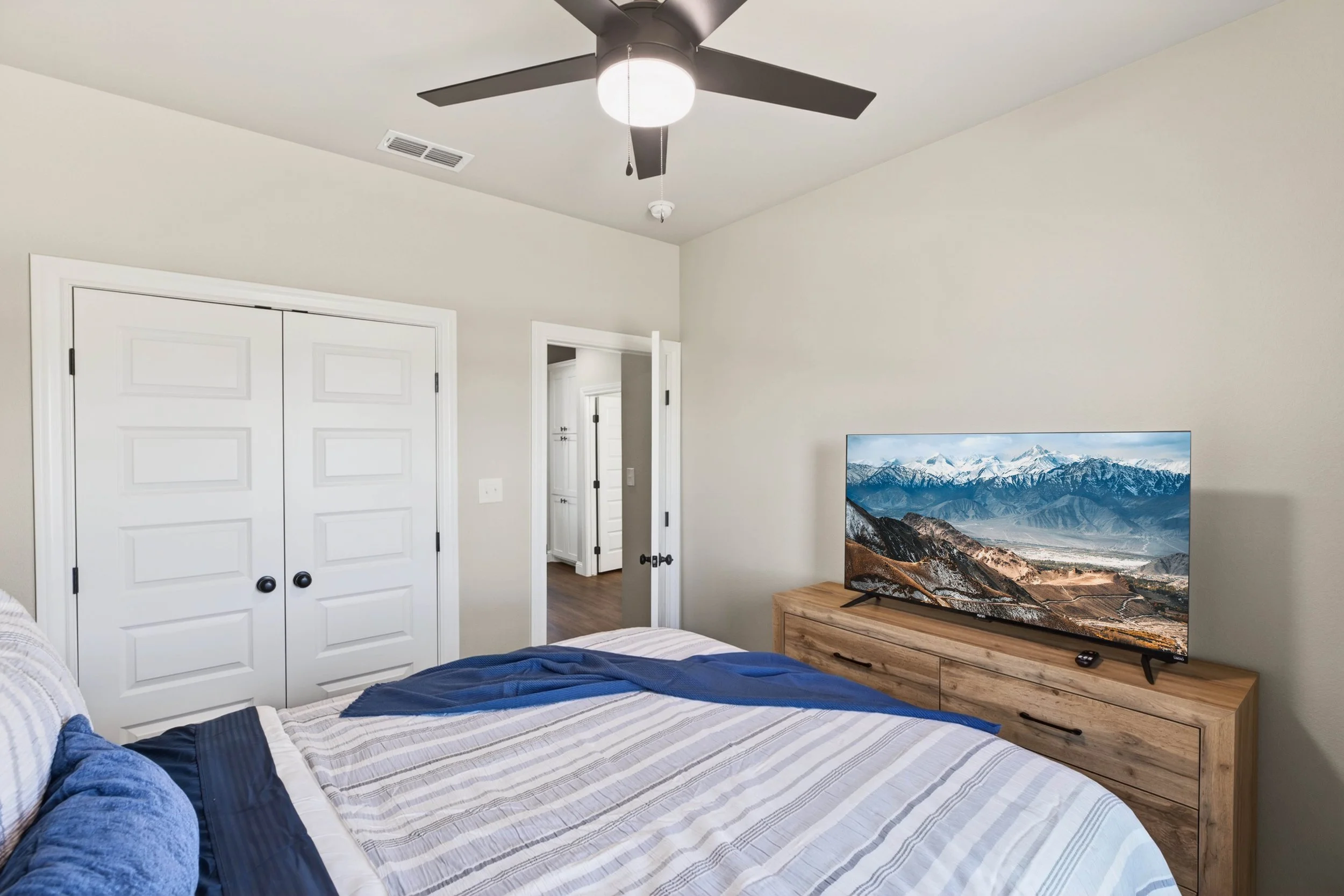 A bedroom with a ceiling fan, a TV showing mountain scenery, a command strip, hardwood floors, white closet doors, and an open doorway leading to another room.