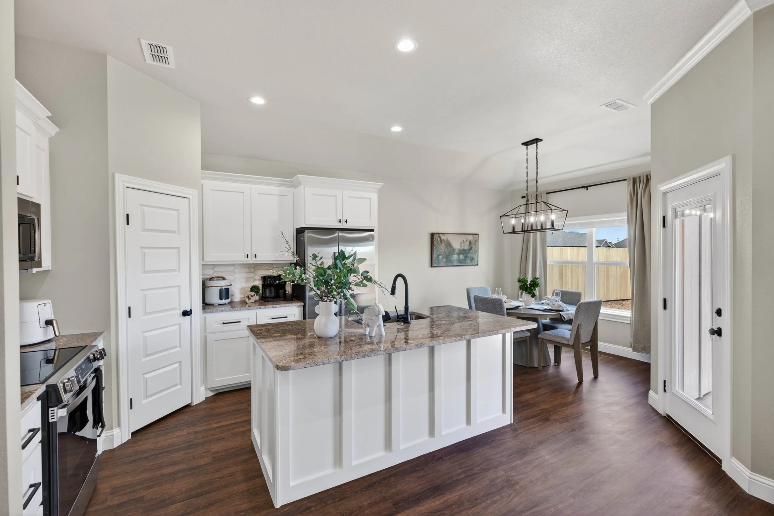 Open-concept kitchen with white cabinets, a kitchen island with a granite countertop, and a dining area with a window and window curtains. There are plants on the dining table, and a chandelier hangs above it. The floor is wooden, and the walls are p