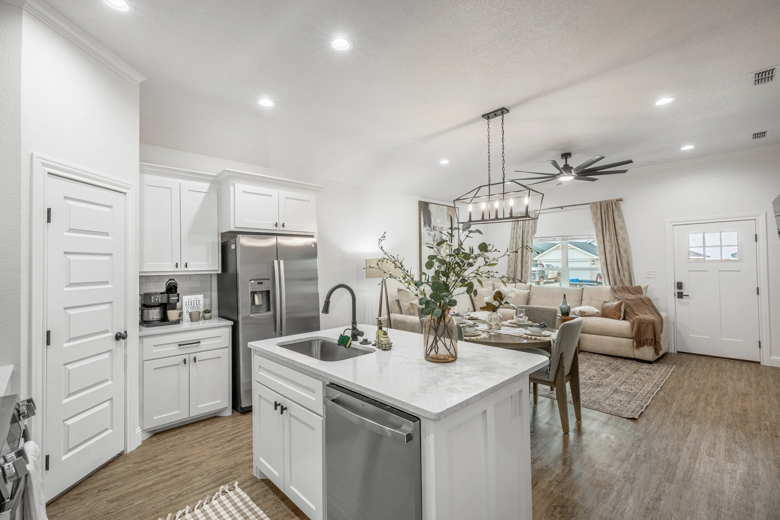 Open-concept kitchen with white cabinets, stainless steel refrigerator, white island with sink, and adjacent living room with a beige sofa, dining table, and decorative items, illuminated by ceiling lights and a chandelier.