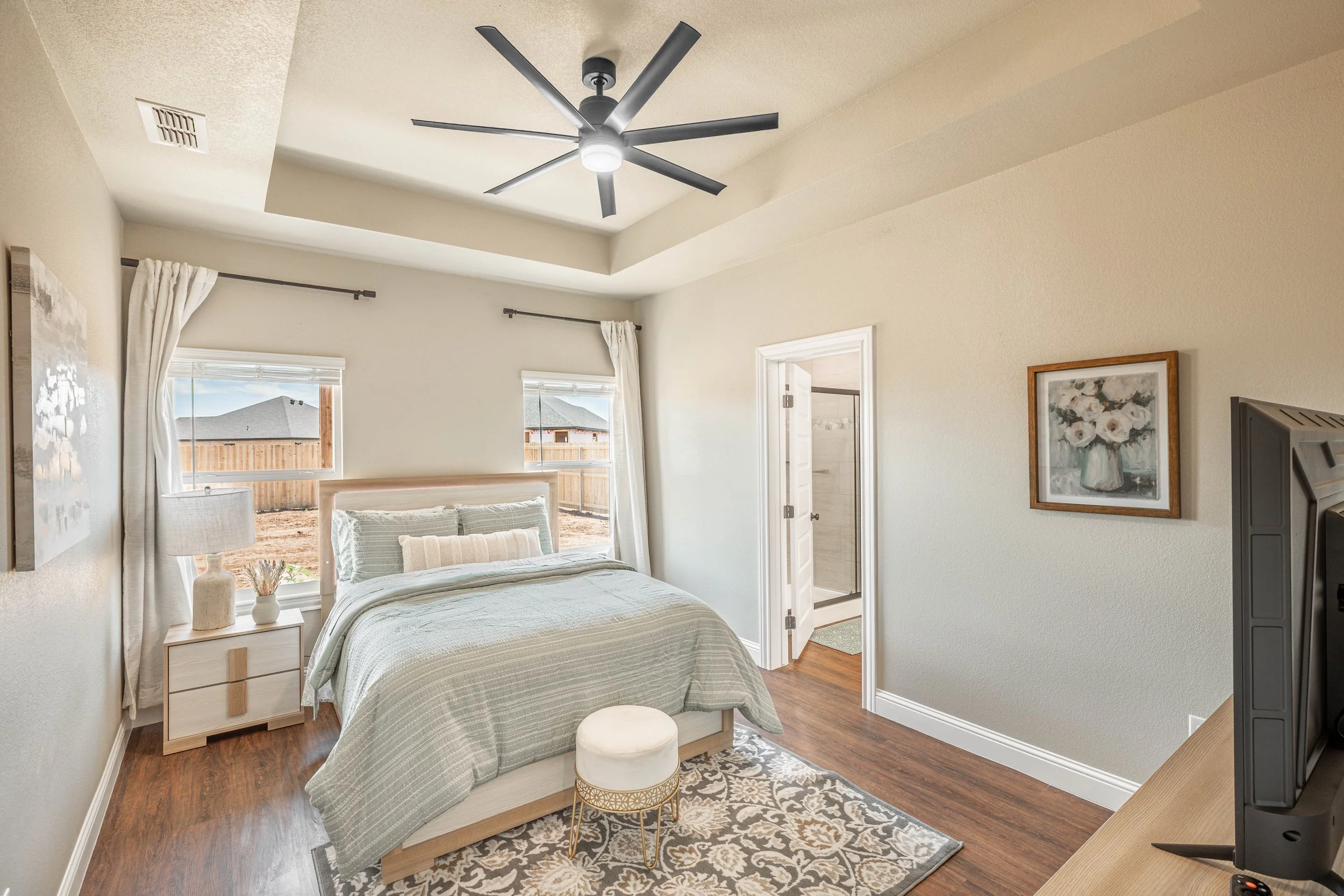 A bedroom with a bed, bedside table, lamp, and a window with curtains, beige walls, wooden floor, ceiling fan, and a picture frame on the wall.