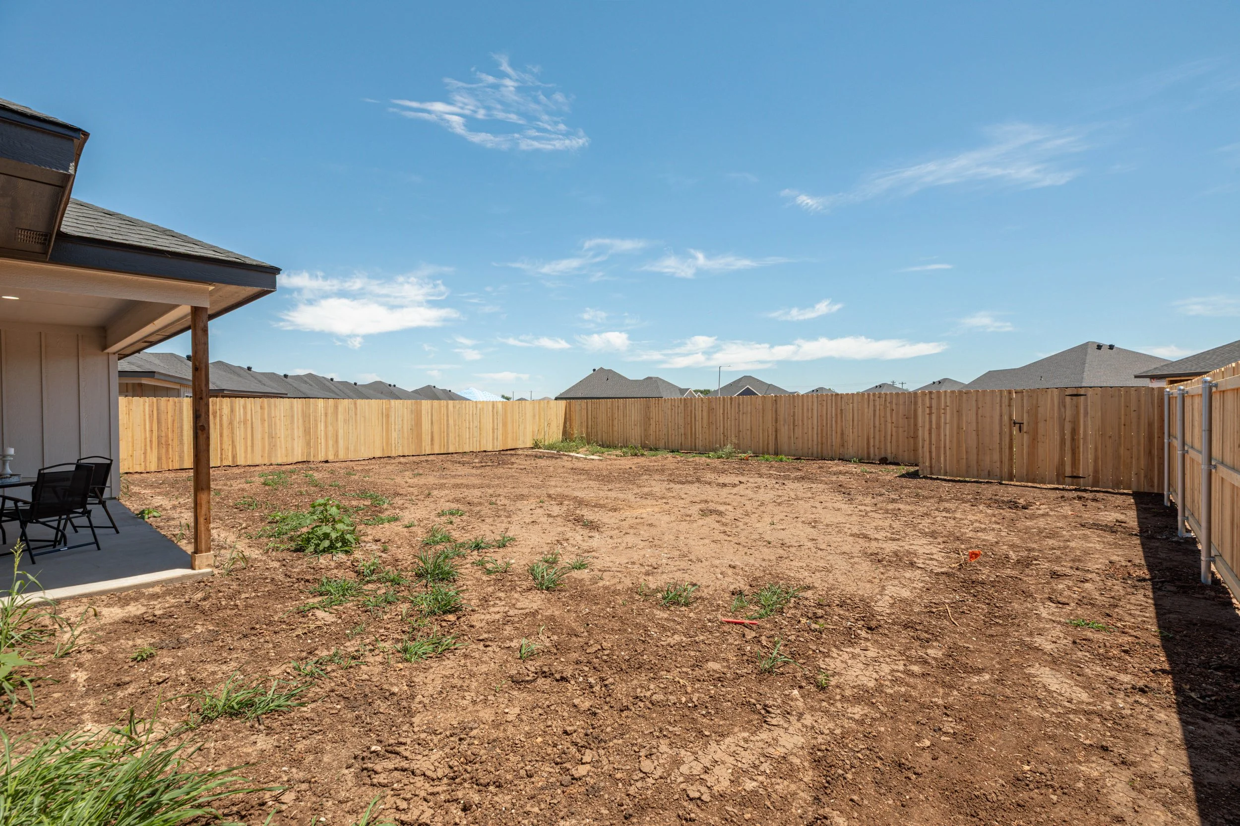 A backyard with a dirt ground and small green plants, enclosed by a wooden fence. Part of a house's porch with chairs and a table is visible on the left. The sky is clear with a few scattered clouds.