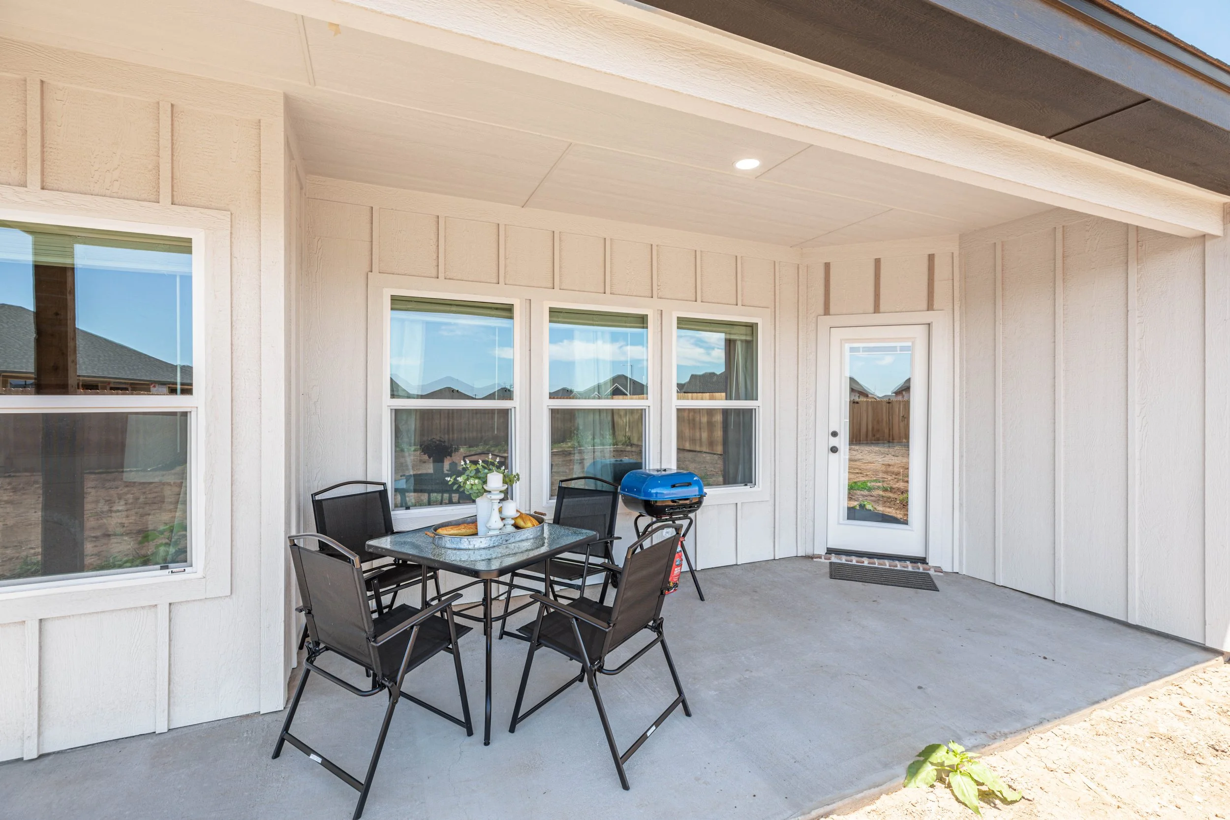 Back porch of a house with four windows and a glass door, a patio table with four chairs, a small grill, and some indoor plants. The porch has a grey floor and is partially covered with a roof.