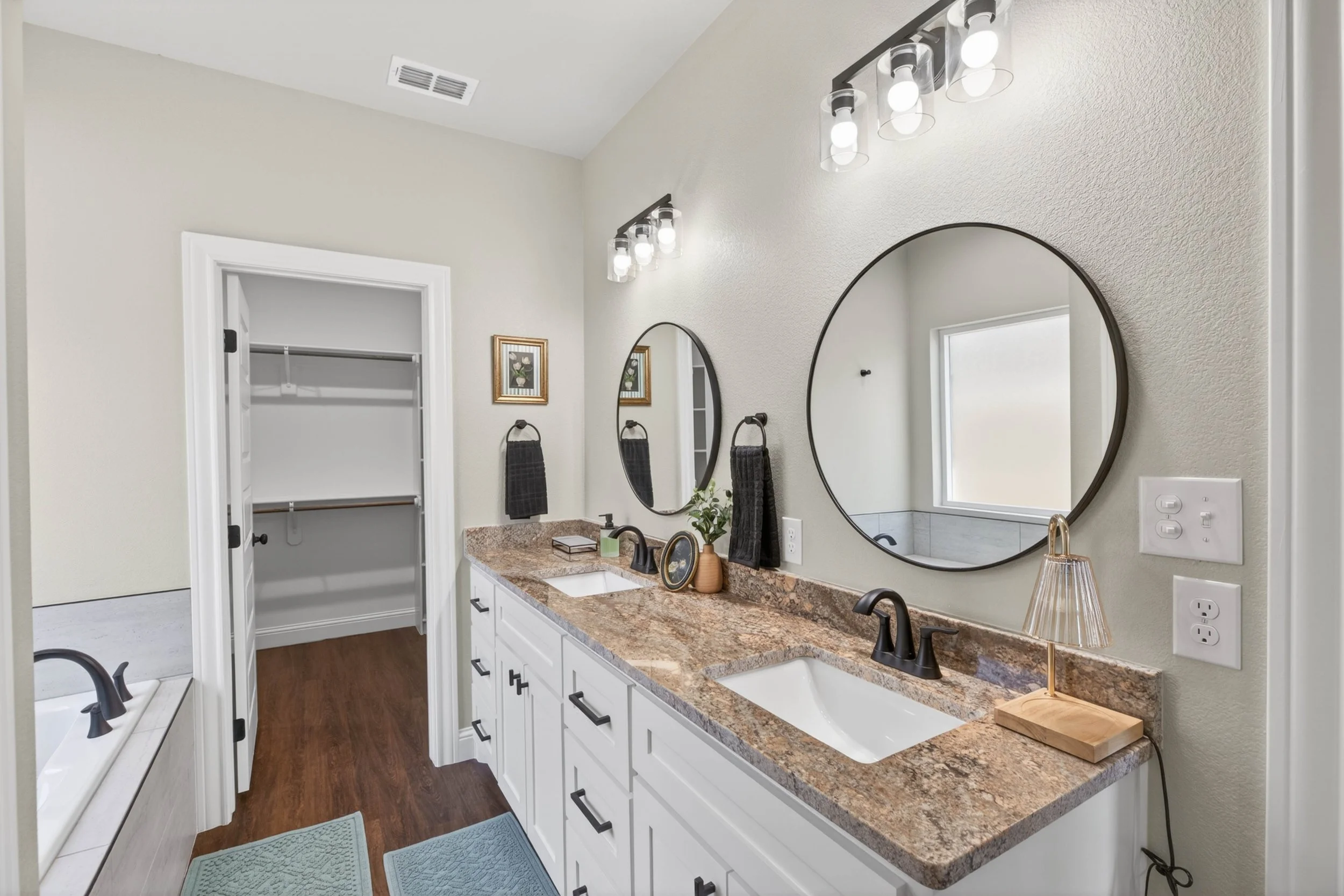 Bathroom with double sinks, large mirrors, light fixtures, framed wall art, towel holders, and a vanity with decorative items, with a closet visible through an open door.