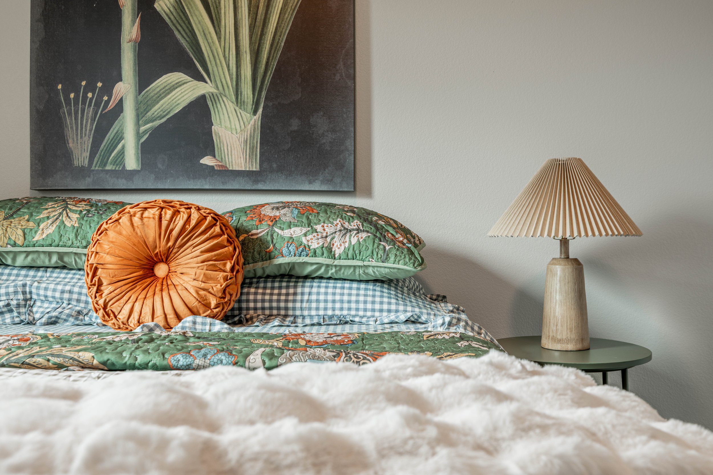 A neatly made bed with a green floral quilt, two patterned pillows, and a large orange round pillow. To the right of the bed is a beige table lamp with a pleated shade on a white side table. On the wall above the bed is a framed botanical print of la