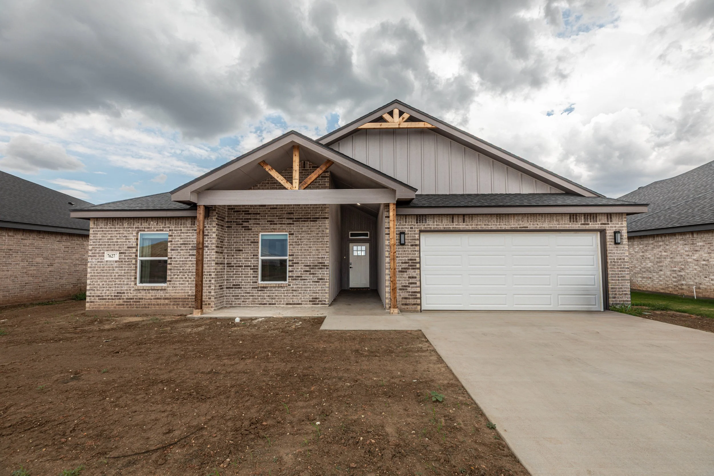Newly constructed brick house with a white garage door and front door, unfinished yard with dirt and a concrete sidewalk, cloudy sky in the background.