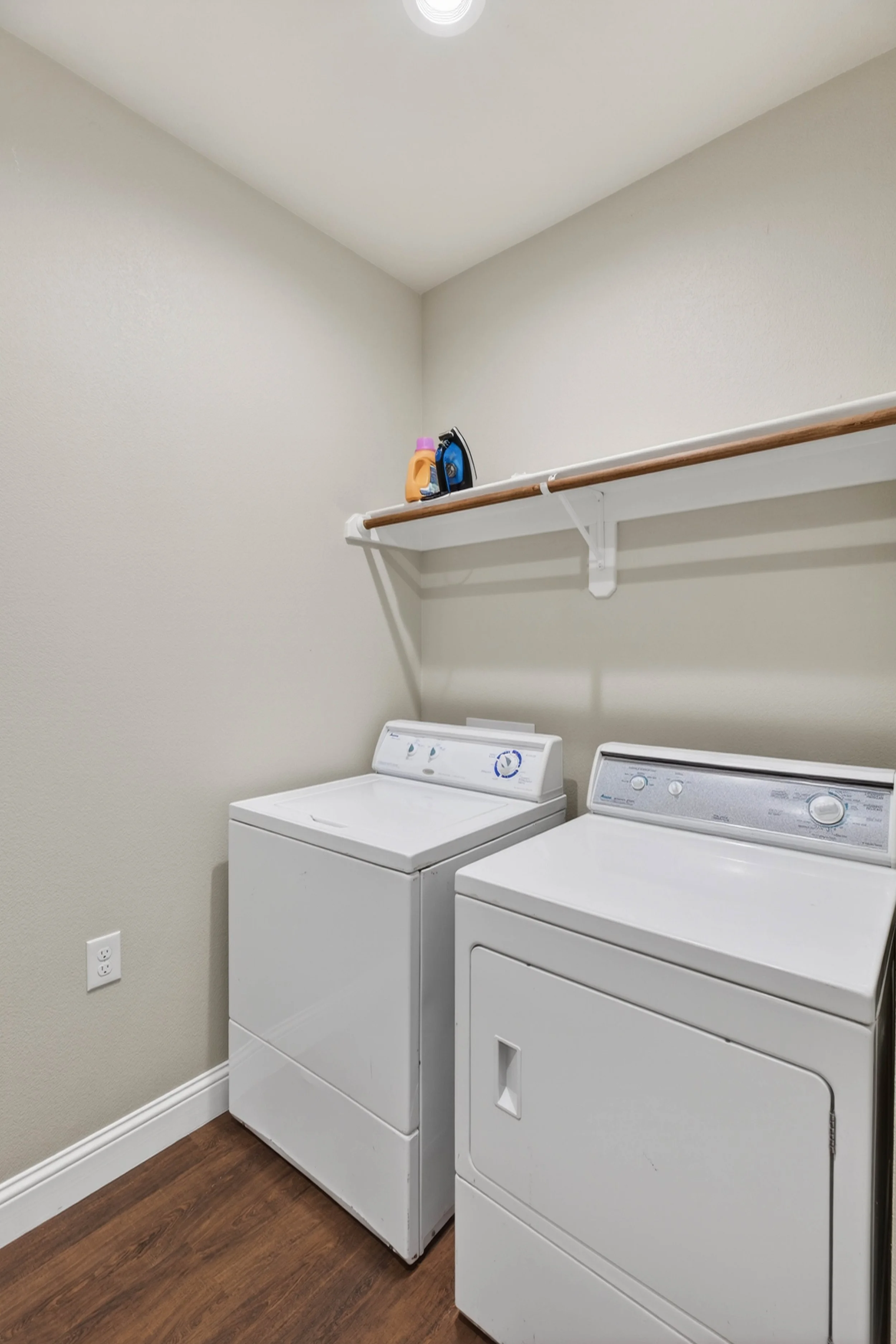 A laundry room with a white washing machine and dryer side by side on a wooden floor. There is a white wall with a wooden shelf above the appliances, holding a bottle of detergent and an iron.