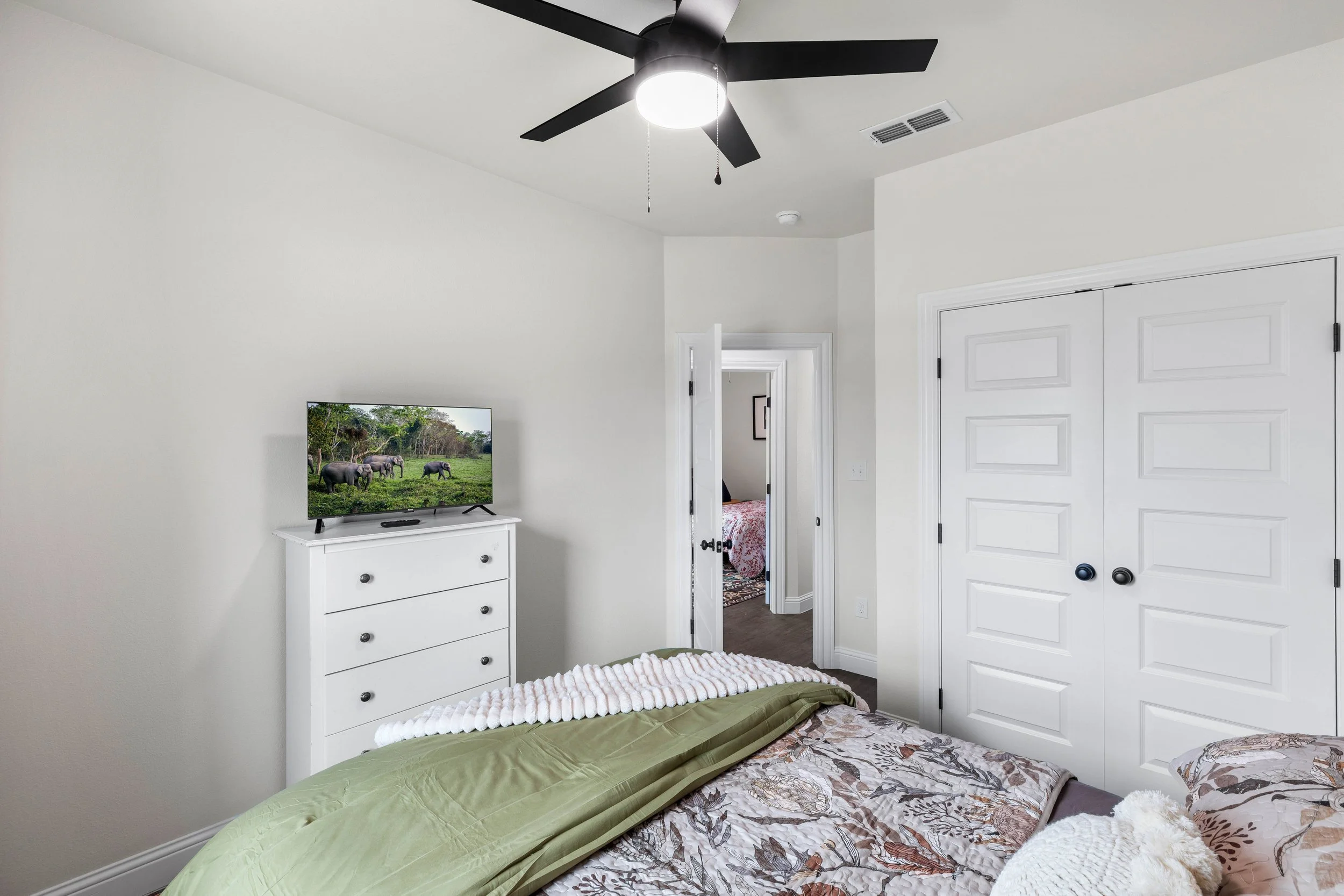 A bedroom with a ceiling fan, white walls, a white dresser with a TV showing elephants, and a partially visible bed with brown, white, and green bedding. There is a door leading to another room and a closet with closed white doors.