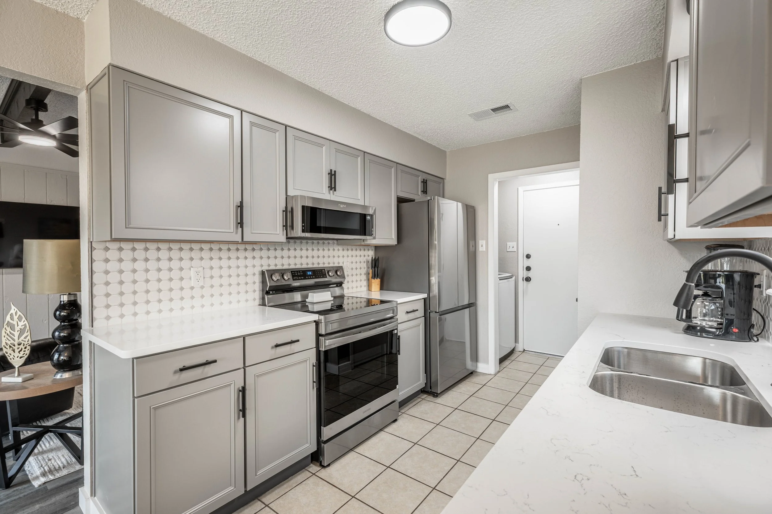 A modern kitchen with gray cabinets, stainless steel appliances including a microwave and refrigerator, a white countertop with a double stainless steel sink, and a hexagonal tile backsplash. There is a small section of a dining area with a table and