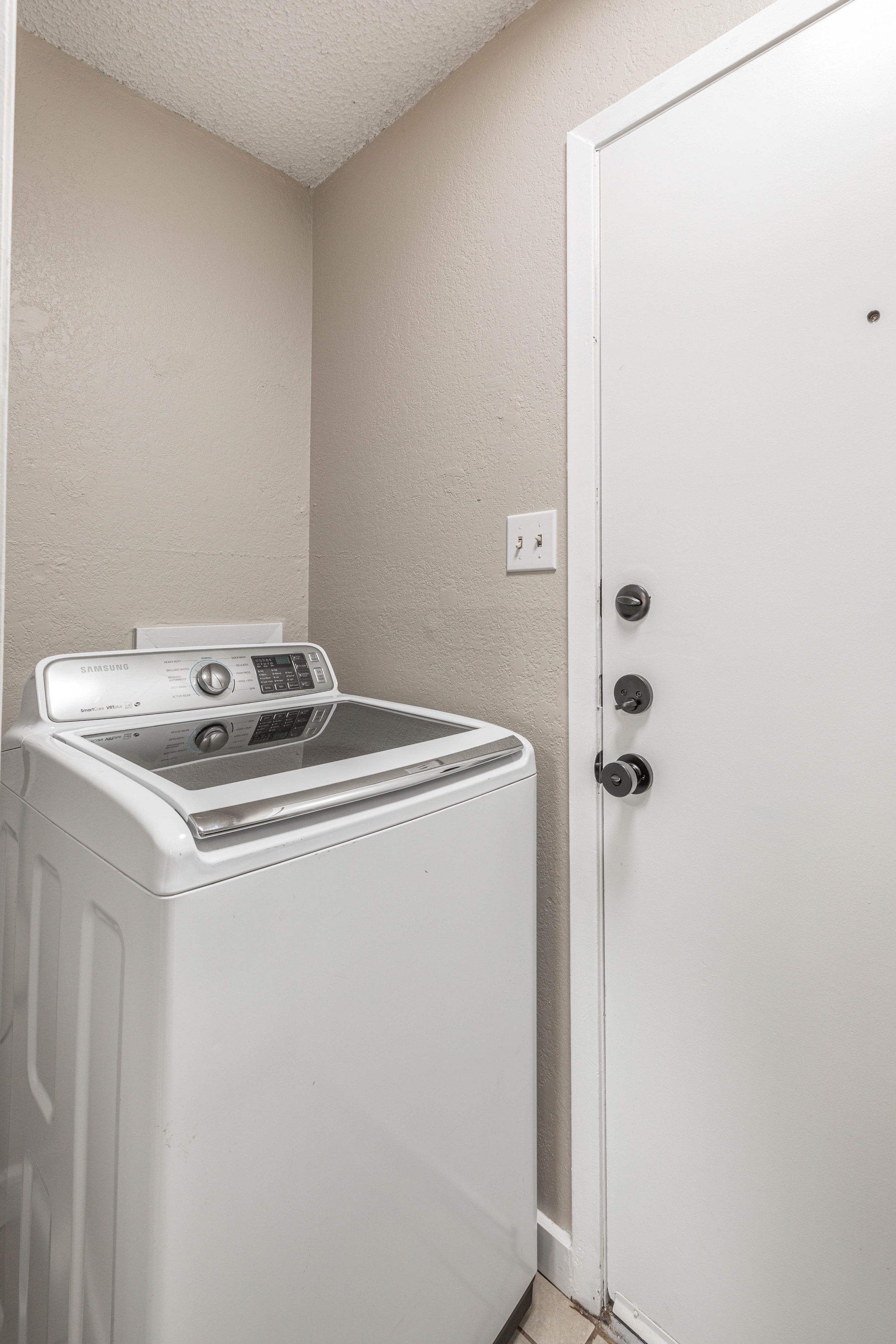 A white Samsung top-loading washing machine with a digital control panel, situated against beige textured walls and a white door with a black doorknob.