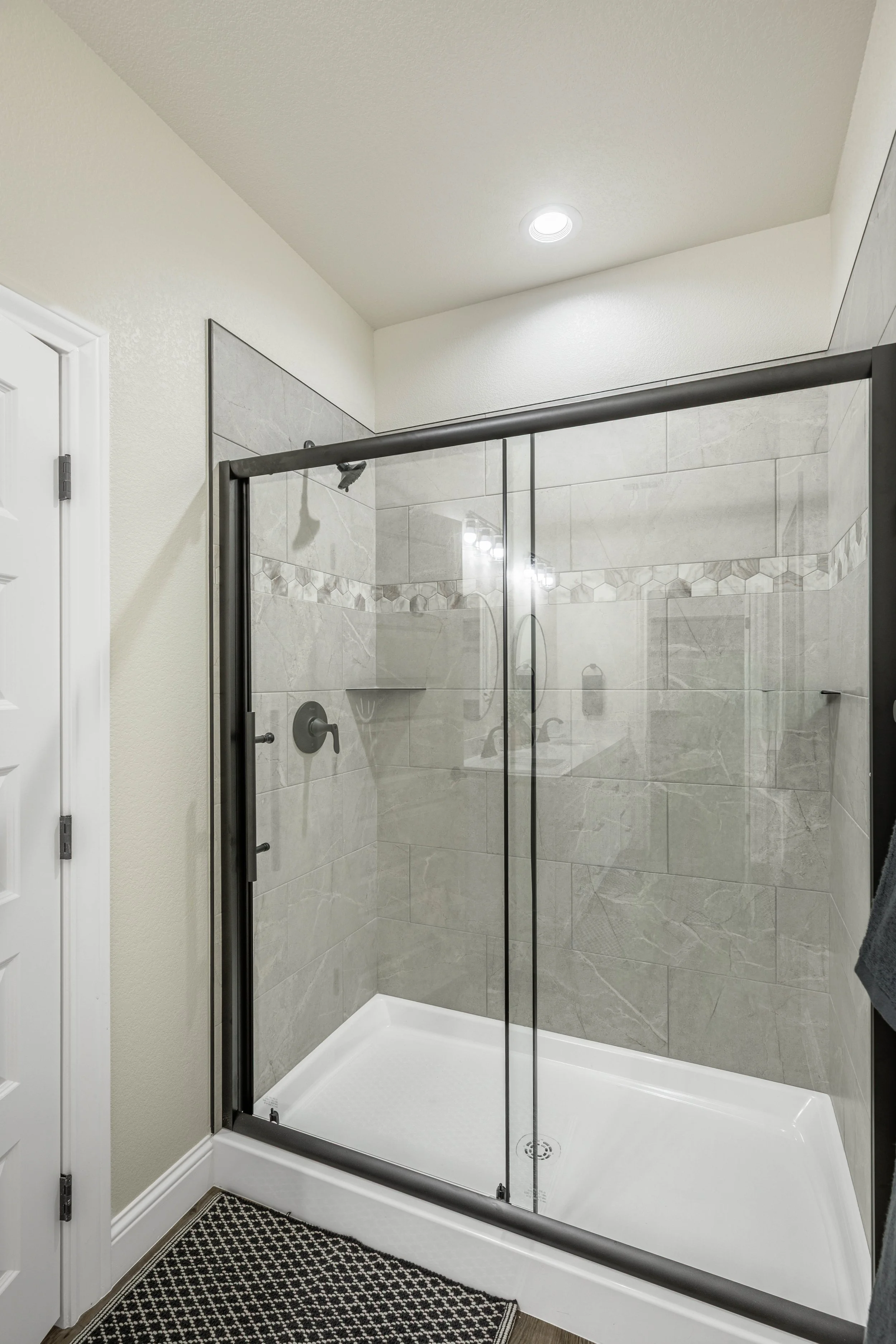 A walk-in shower with a black metal frame, large beige marble tiles with a decorative border, and a white shower base. Part of the bathroom with a white door and a black and white patterned rug.