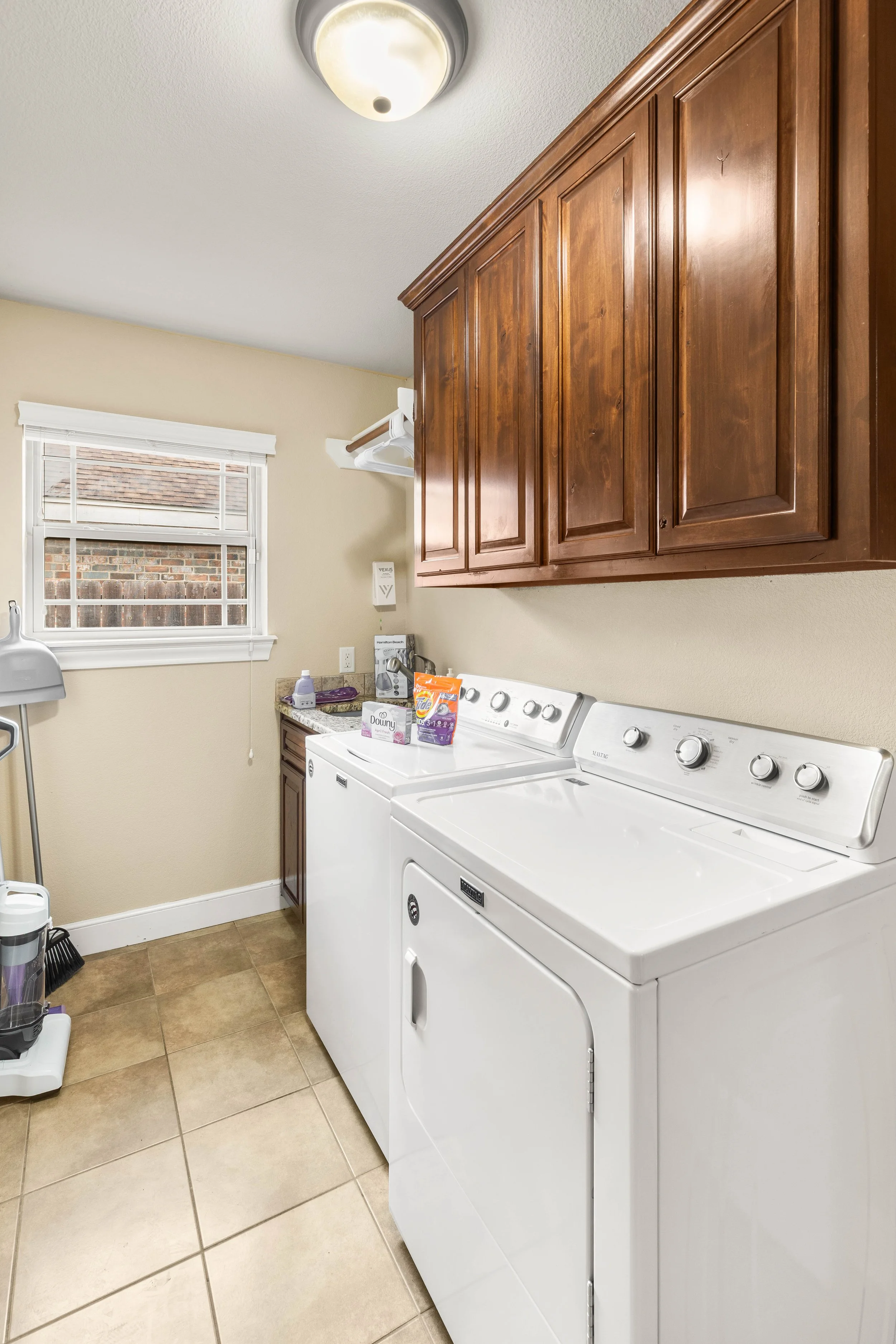 A laundry room with a white washer and dryer side by side, wooden cabinets above, a window with blinds, and various laundry supplies on top.