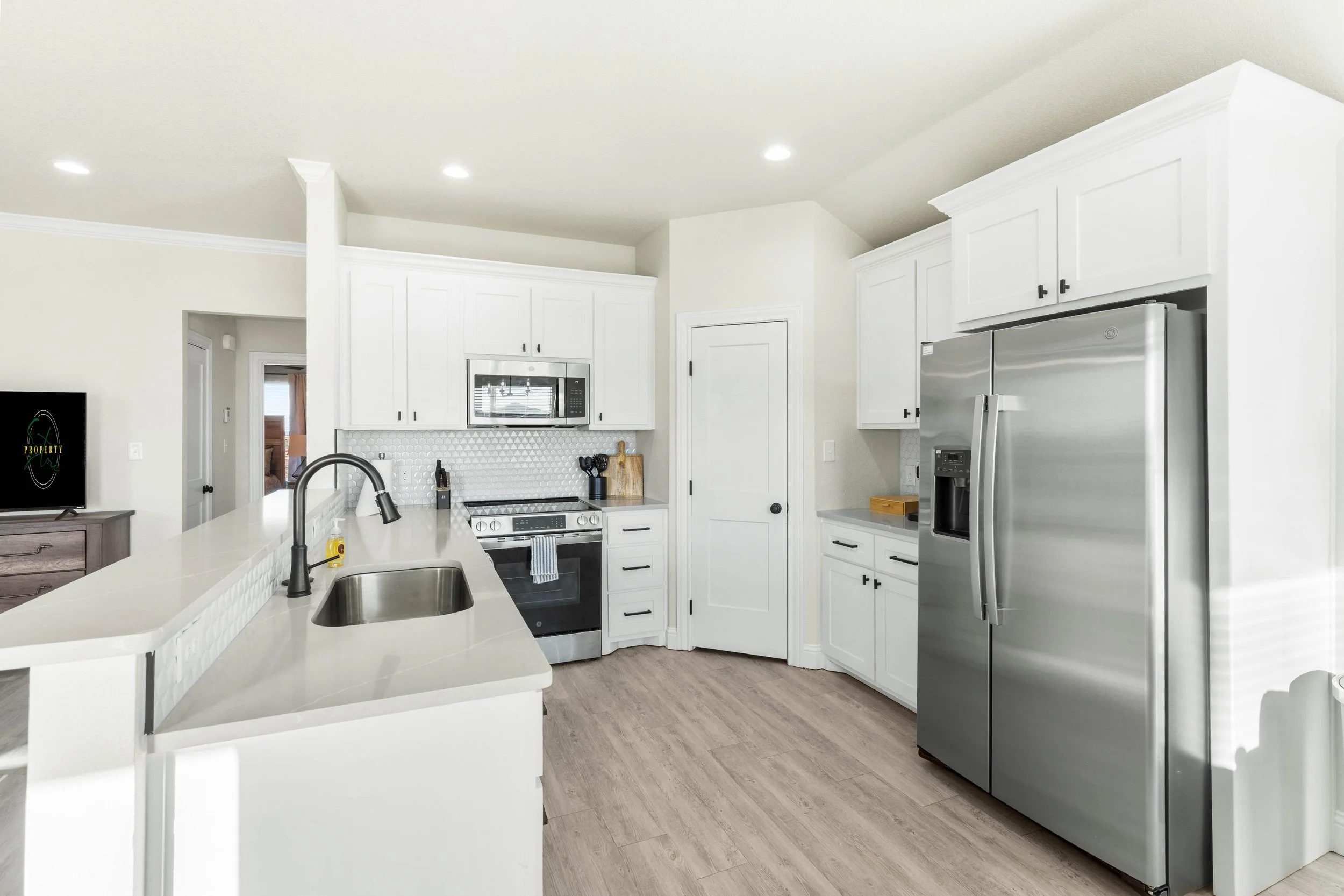 Modern white kitchen with stainless steel appliances, including a side-by-side refrigerator and microwave, white cabinets, a white countertop island with a sink, and wood-like flooring.