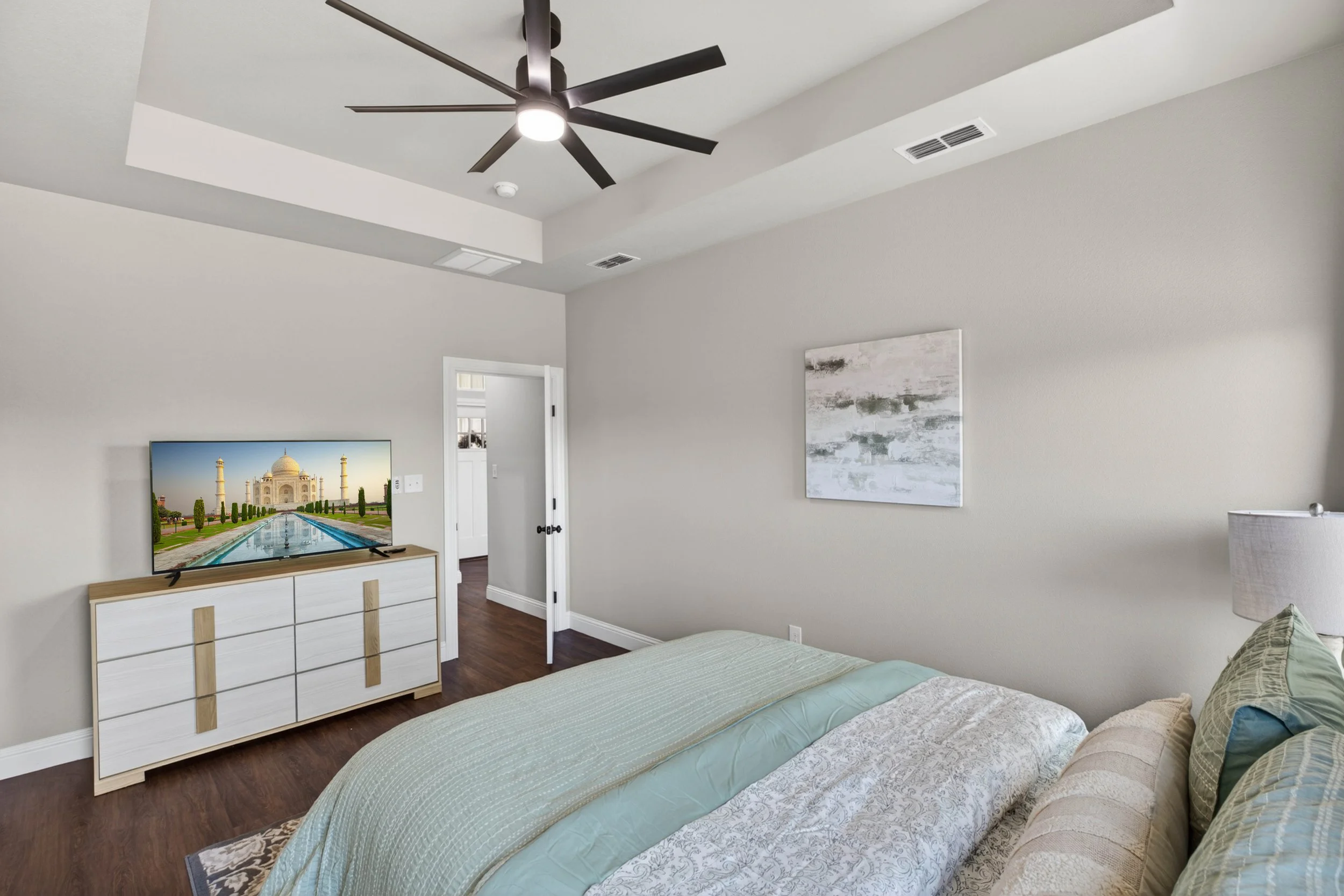 A bedroom with a bed, dresser with a TV displaying the Taj Mahal, wall art, and a ceiling fan.