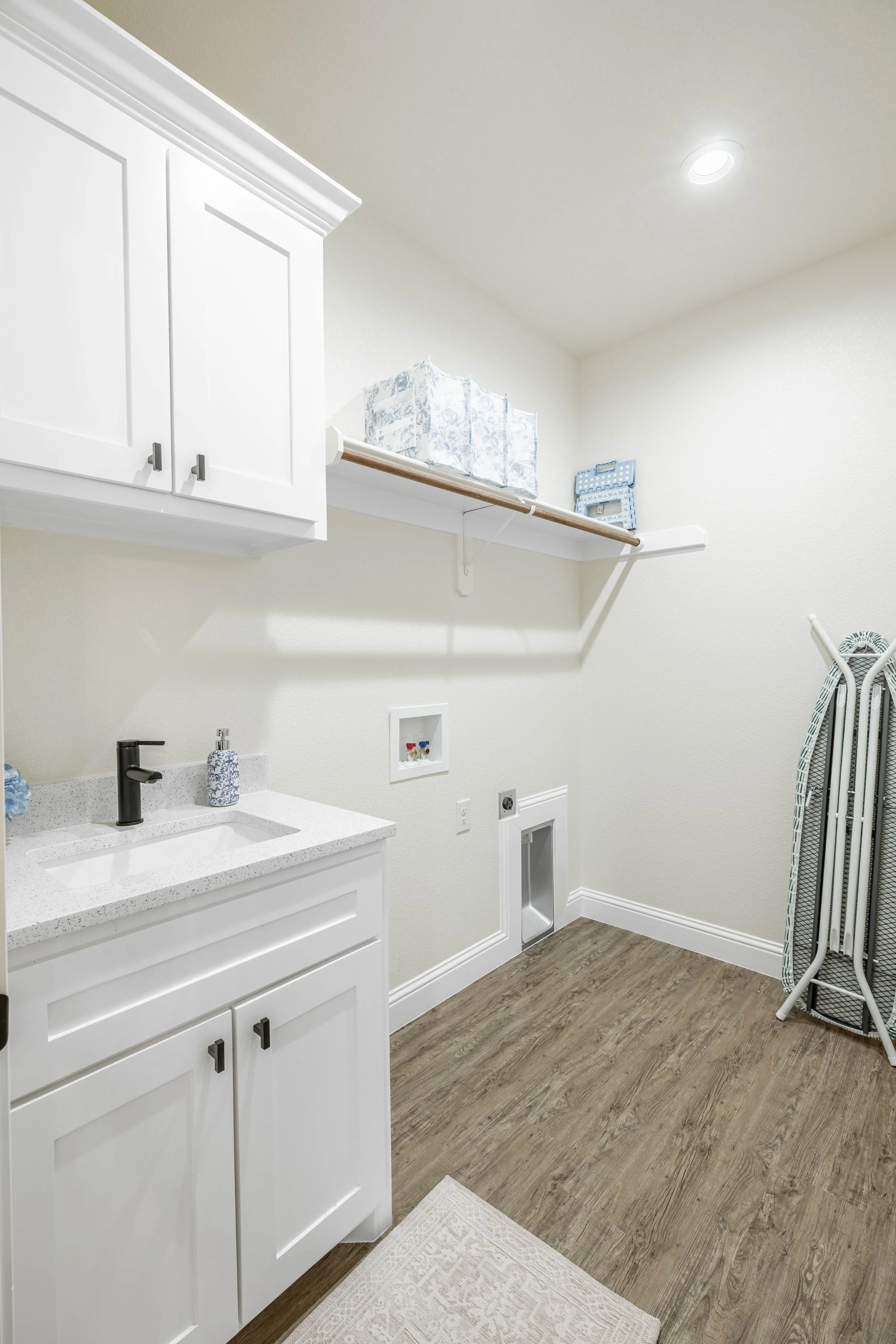 Laundry room with white cabinetry, a small sink, and an ironing board next to the wall. There is a shelf with storage boxes, a soap dispenser, a laundry outlet, and a small doorway for utilities.