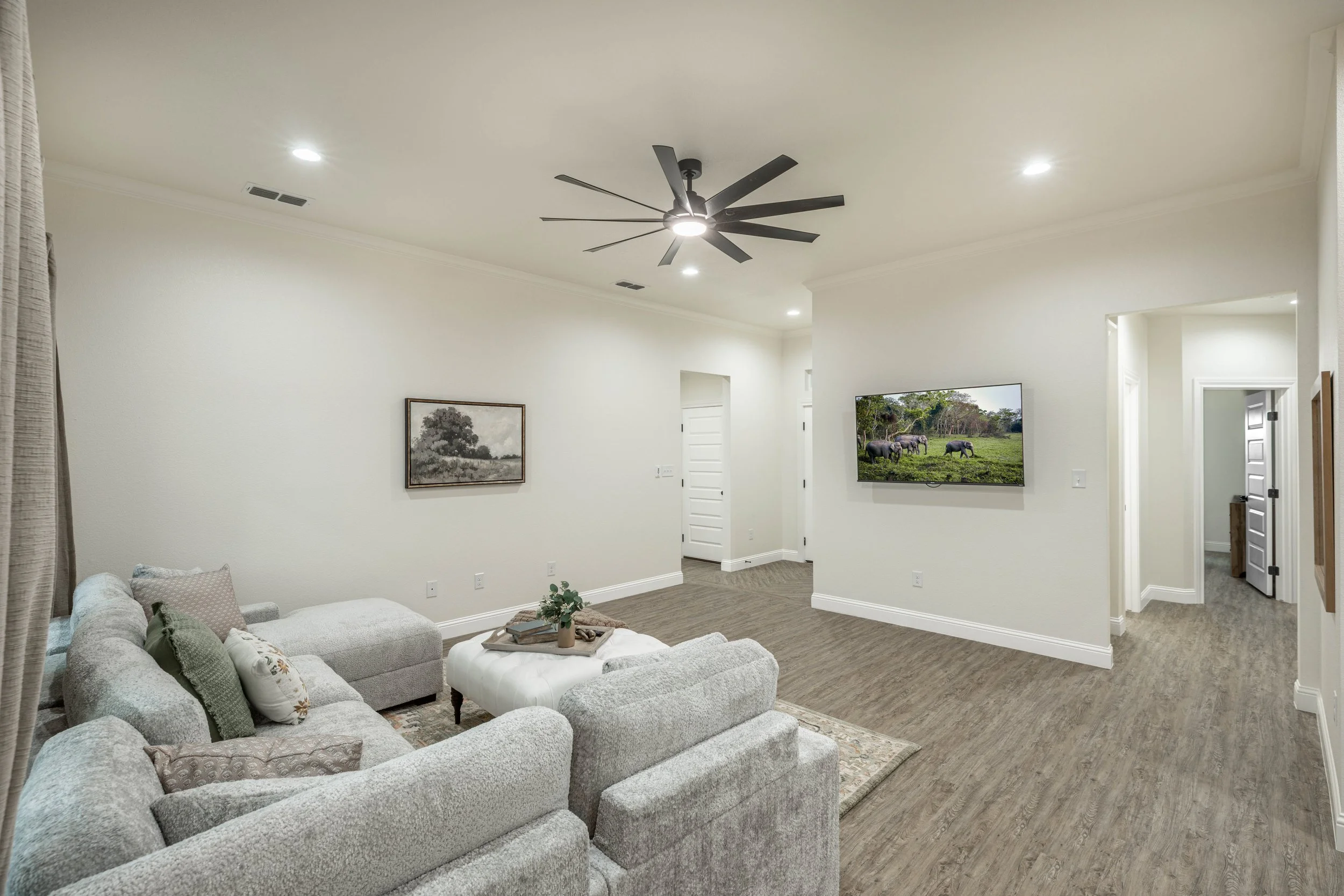 Living room with light-colored sectional sofa, a pillow, a small ottoman, a tray with decor, a picture on the wall, a wall-mounted TV showing elephants, and a doorway, with a ceiling fan and recessed lighting.