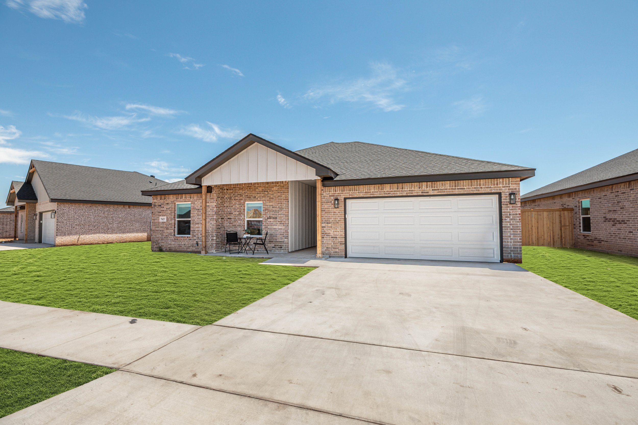 Front view of a modern brick house with a two-car garage, small patio area with outdoor furniture, well-maintained green lawn, and a clear blue sky.