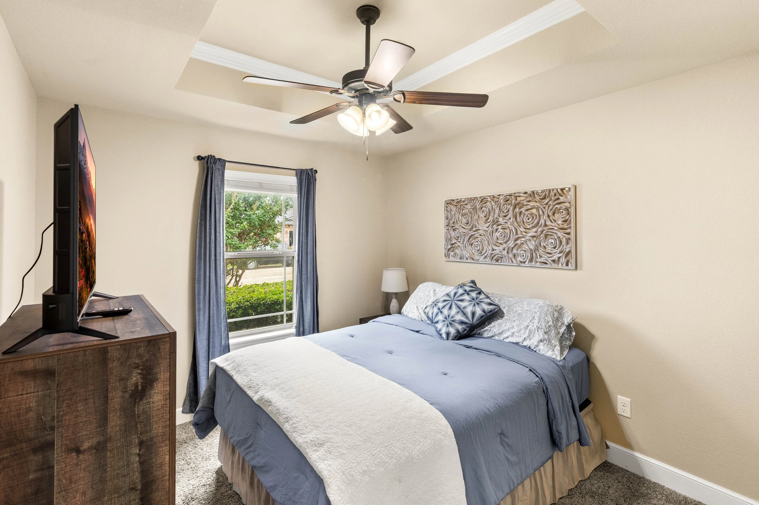 A bedroom with a bed covered in a blue and white quilt, a wooden dresser with a TV, a window with blue curtains, artwork on the wall, and a ceiling fan with lights.
