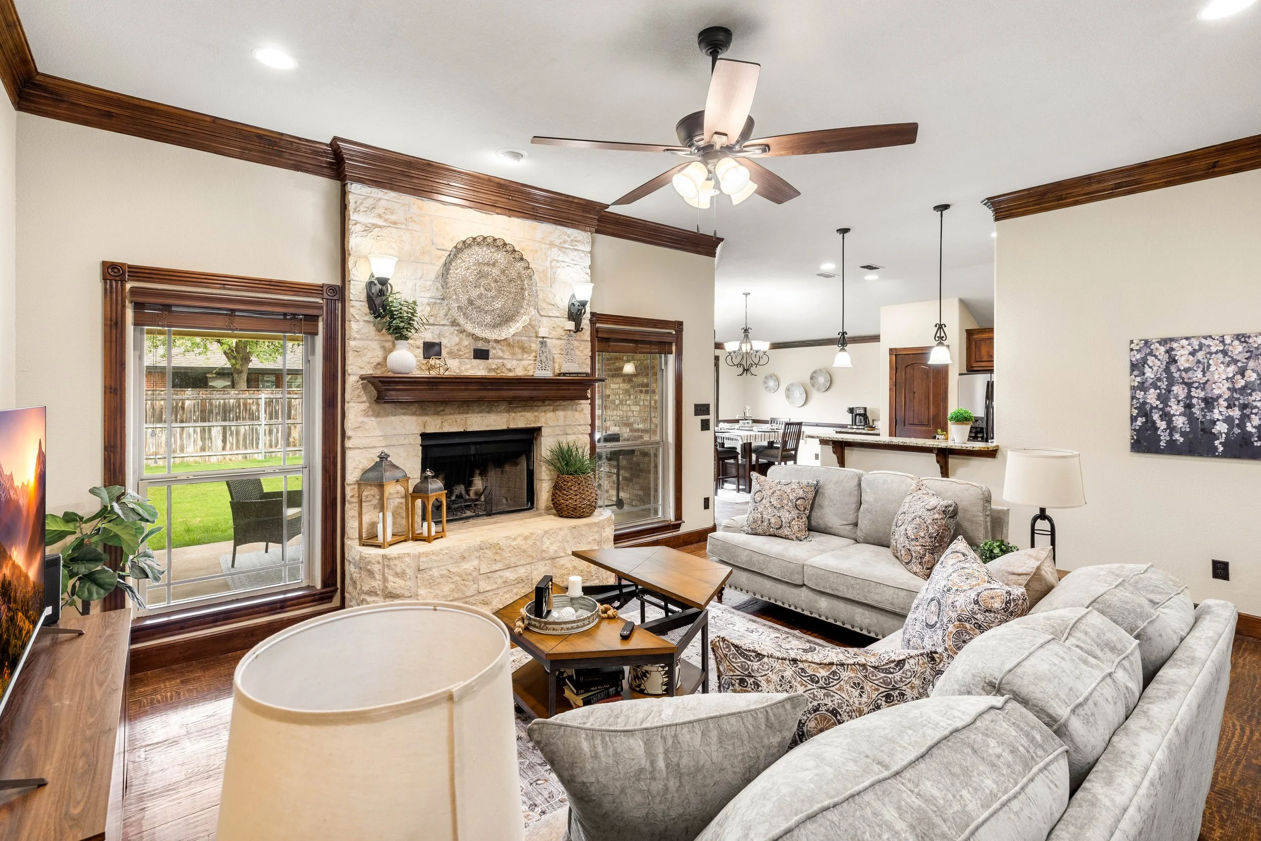 A living room with beige walls, wooden crown molding, a stone fireplace with decorative objects on the mantle, a ceiling fan with lights, a sectional sofa with patterned cushions, a wooden coffee table, and nearby windows and a door leading to an out