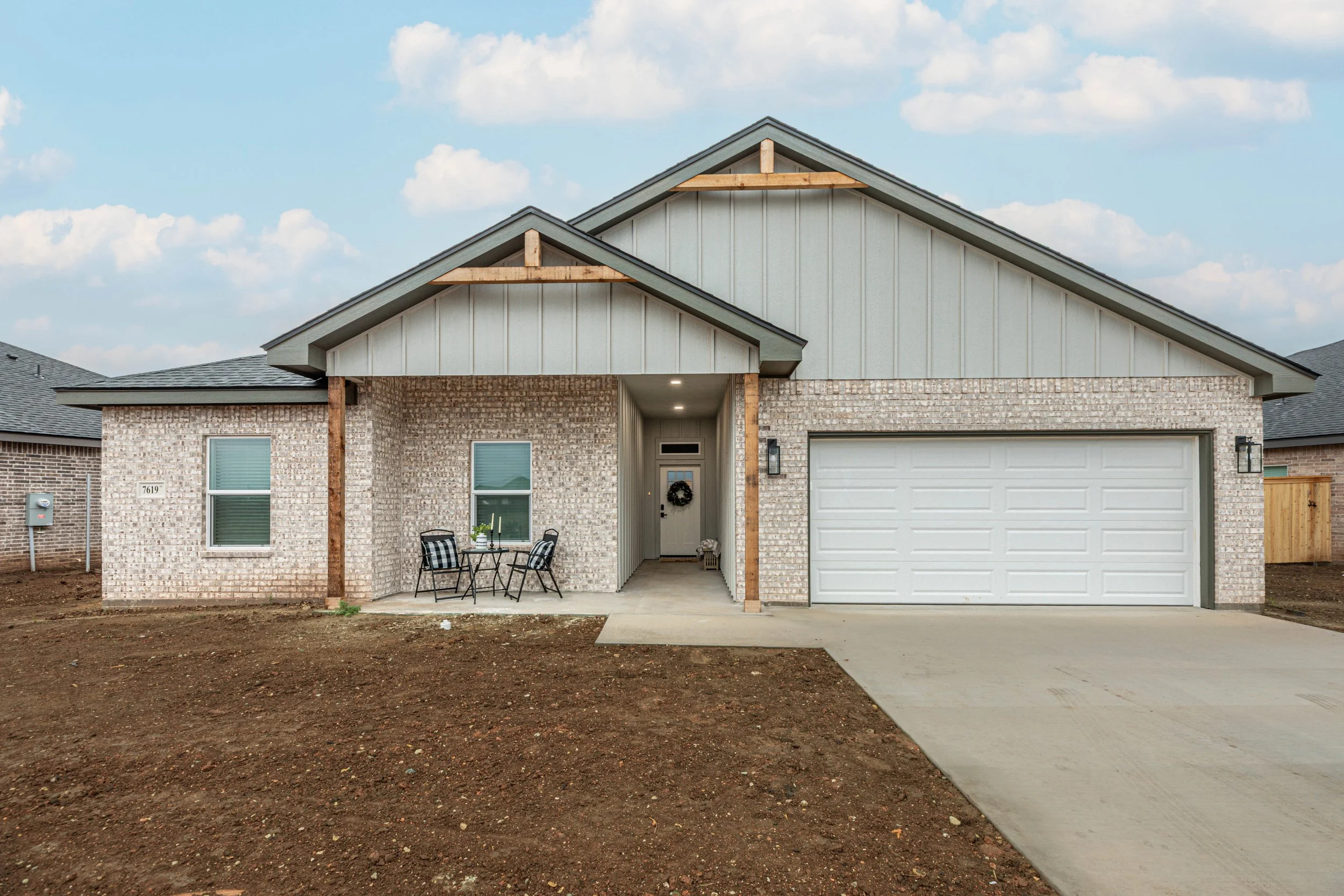 Newly built house with brick exterior and a white garage door, small porch with patio furniture, and a bundle decoration on the front door, under a partly cloudy sky.