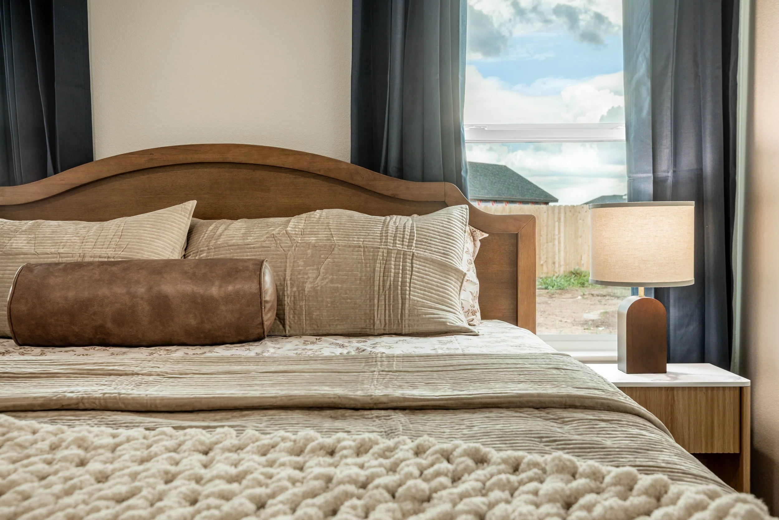 A bedroom with a wooden headboard, beige pillows, a beige blanket, a bedside lamp, and a window with blue curtains showing a view of the sky and neighboring houses.
