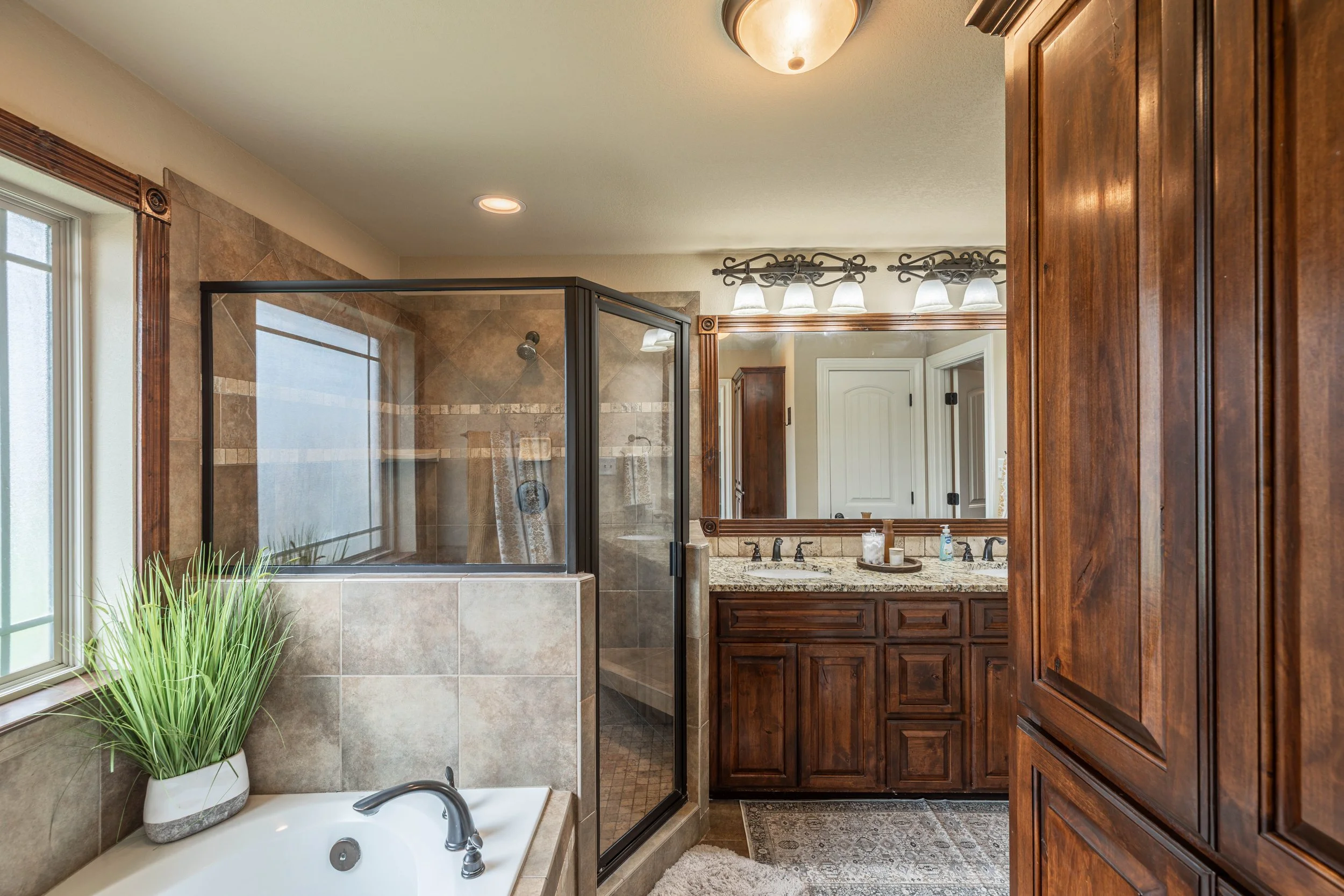 View of a bathroom with a bathtub, shower with glass door, wooden cabinets, granite countertop, and decorative lighting fixtures.
