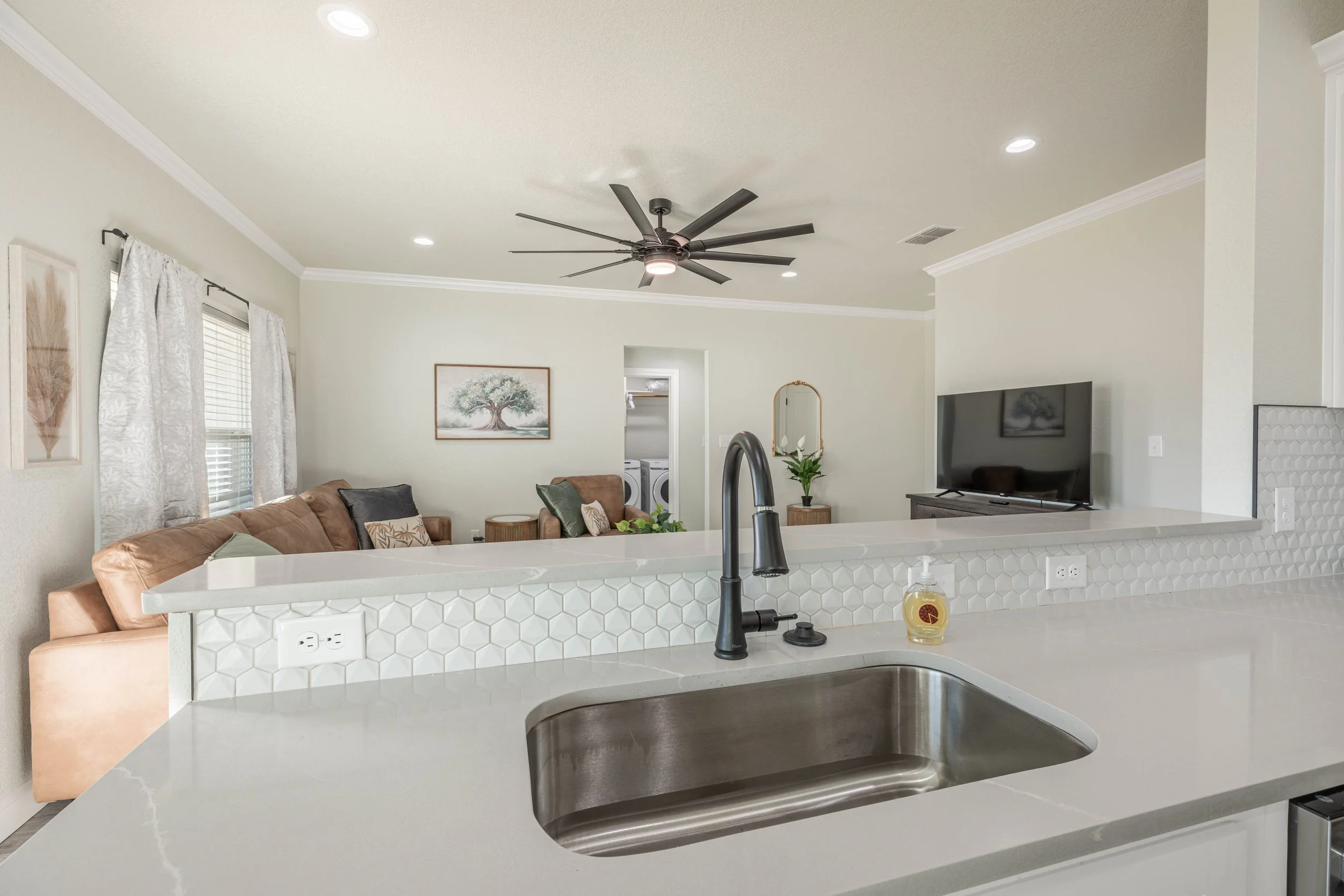 Modern kitchen with white hexagon tile backsplash, black faucet, and open-plan living room with sofa, wall art, TV, and ceiling fan.